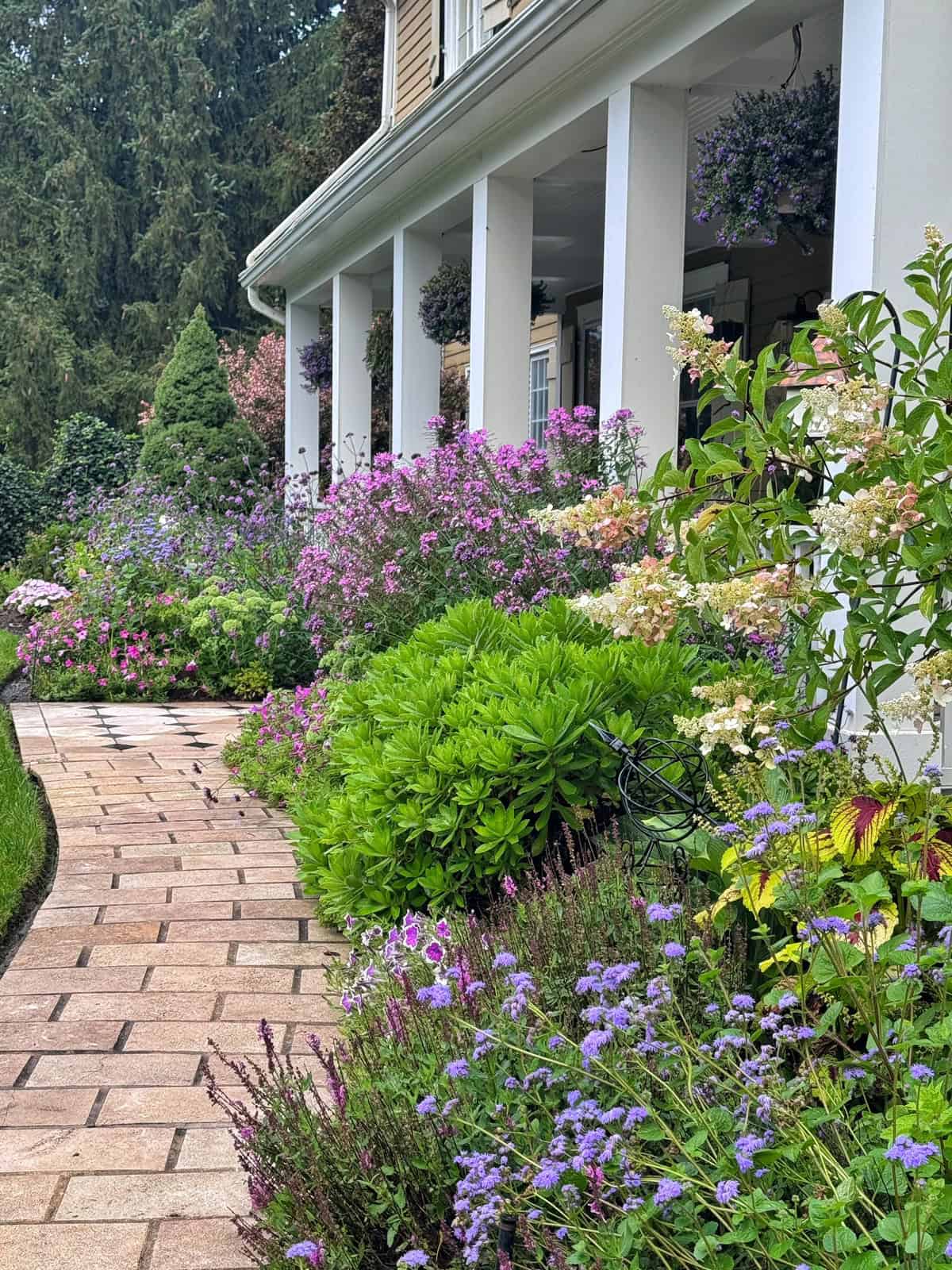 A stone path curves past vibrant, blooming flowers and lush green shrubs alongside a house with white pillars and hanging baskets.