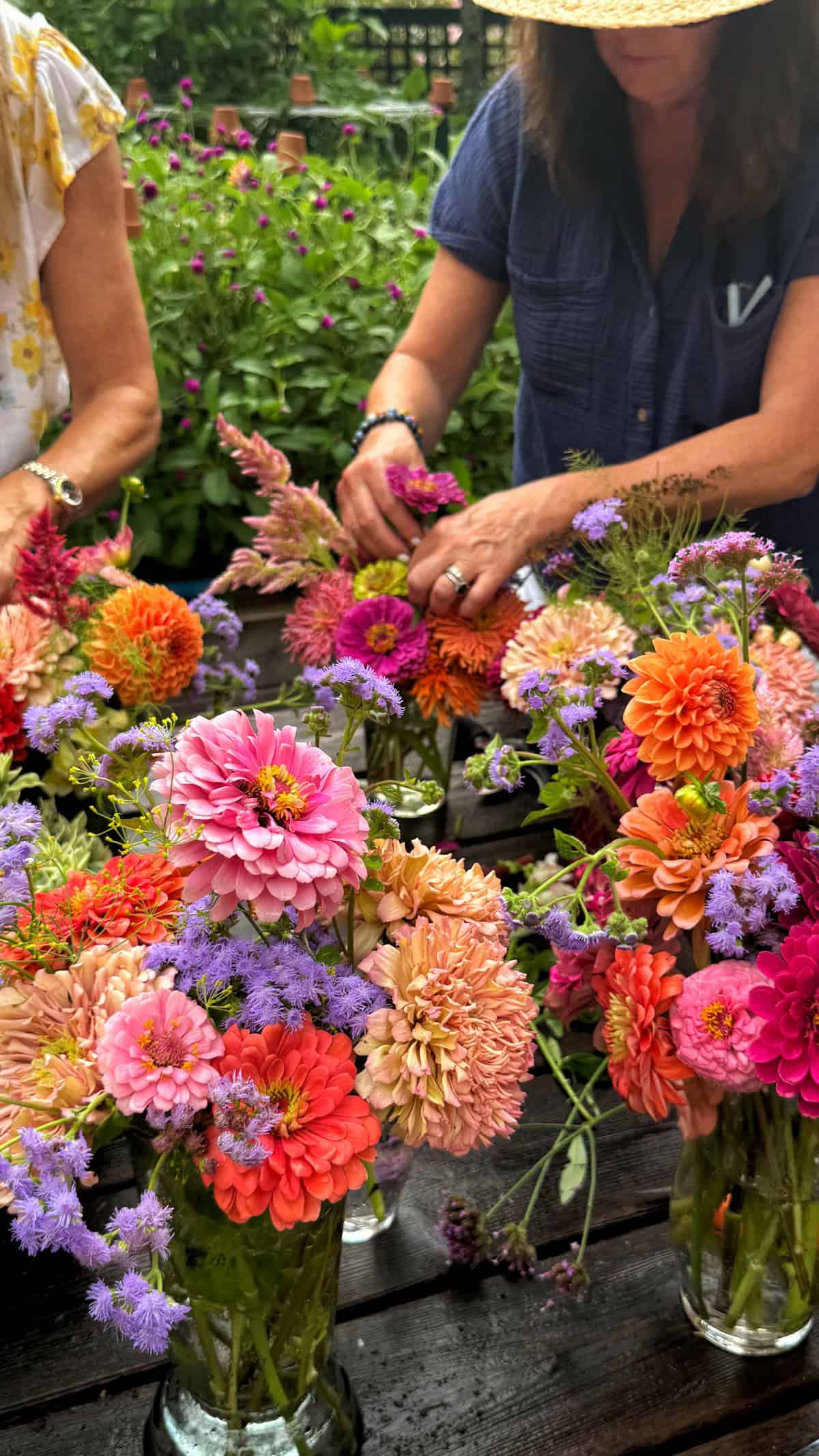 Two women arrange colorful bouquets of zinnias and purple flowers in glass jars on a dark table outdoors, surrounded by green plants.