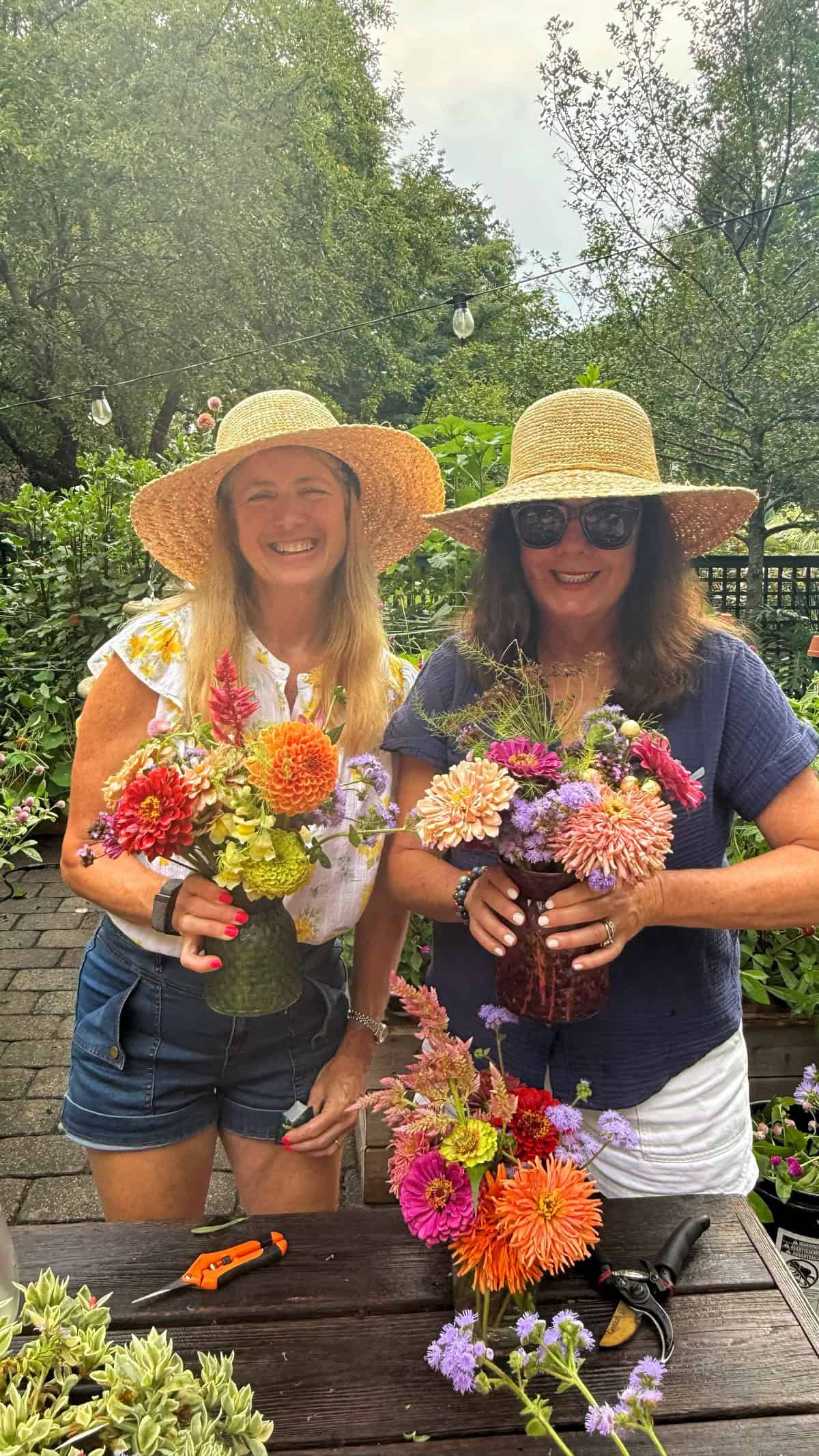 Two women in straw hats smile outdoors, holding colorful bouquets of flowers. They stand at a wooden table with garden tools and flowers, surrounded by lush green trees and plants.