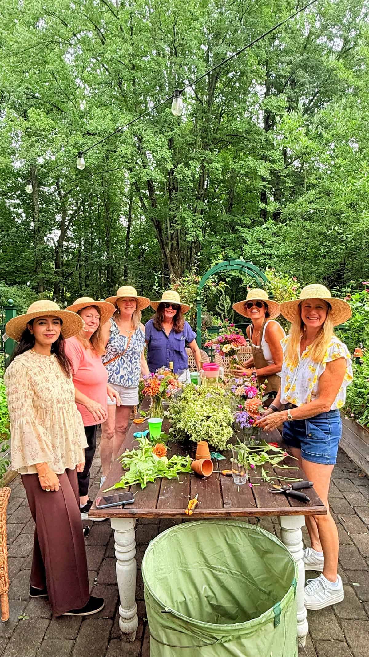 Seven women in summer hats stand around a table with flowers and greenery, smiling outdoors in a lush garden. The table holds bouquets, drinks, and phones. Trees and garden beds surround the group.