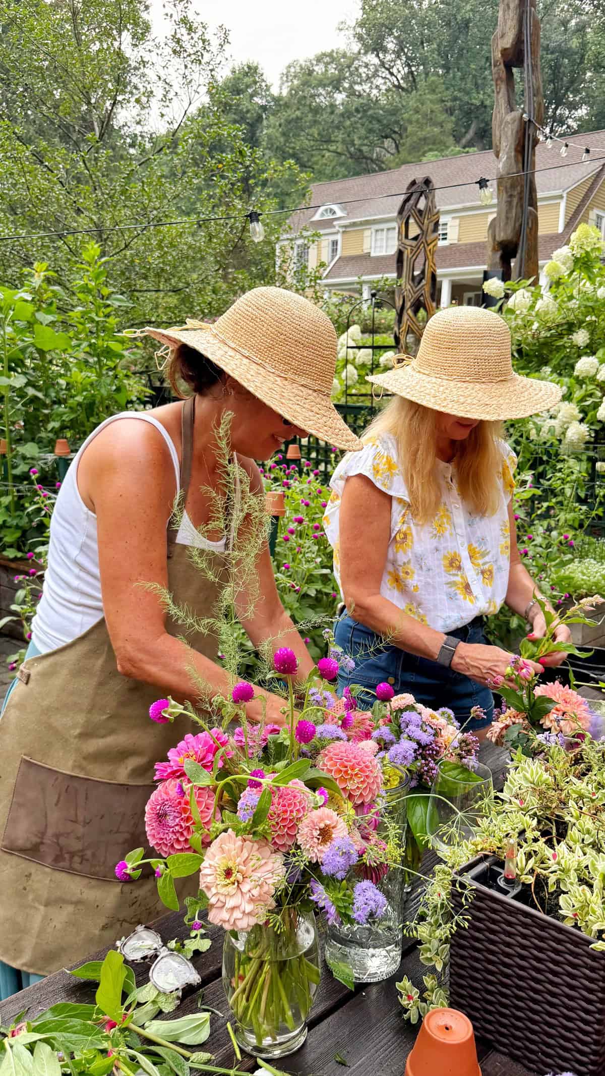Two women in straw hats arrange colorful flowers at an outdoor table in a lush garden. A house and trees are visible in the background. The scene is bright and vibrant, evoking a sense of summer and creativity.