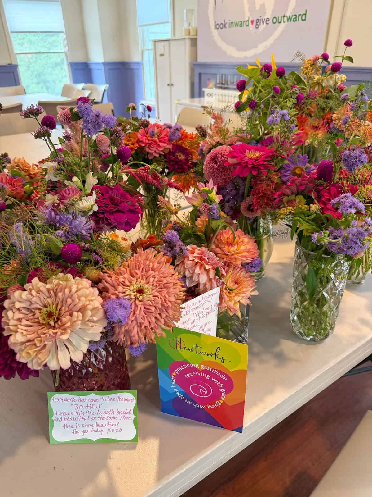 Colorful bouquets of fresh flowers in glass vases sit on a table, with handwritten notes and a Heartworks card among them. In the background, a wall sign reads “look inward give outward.”.