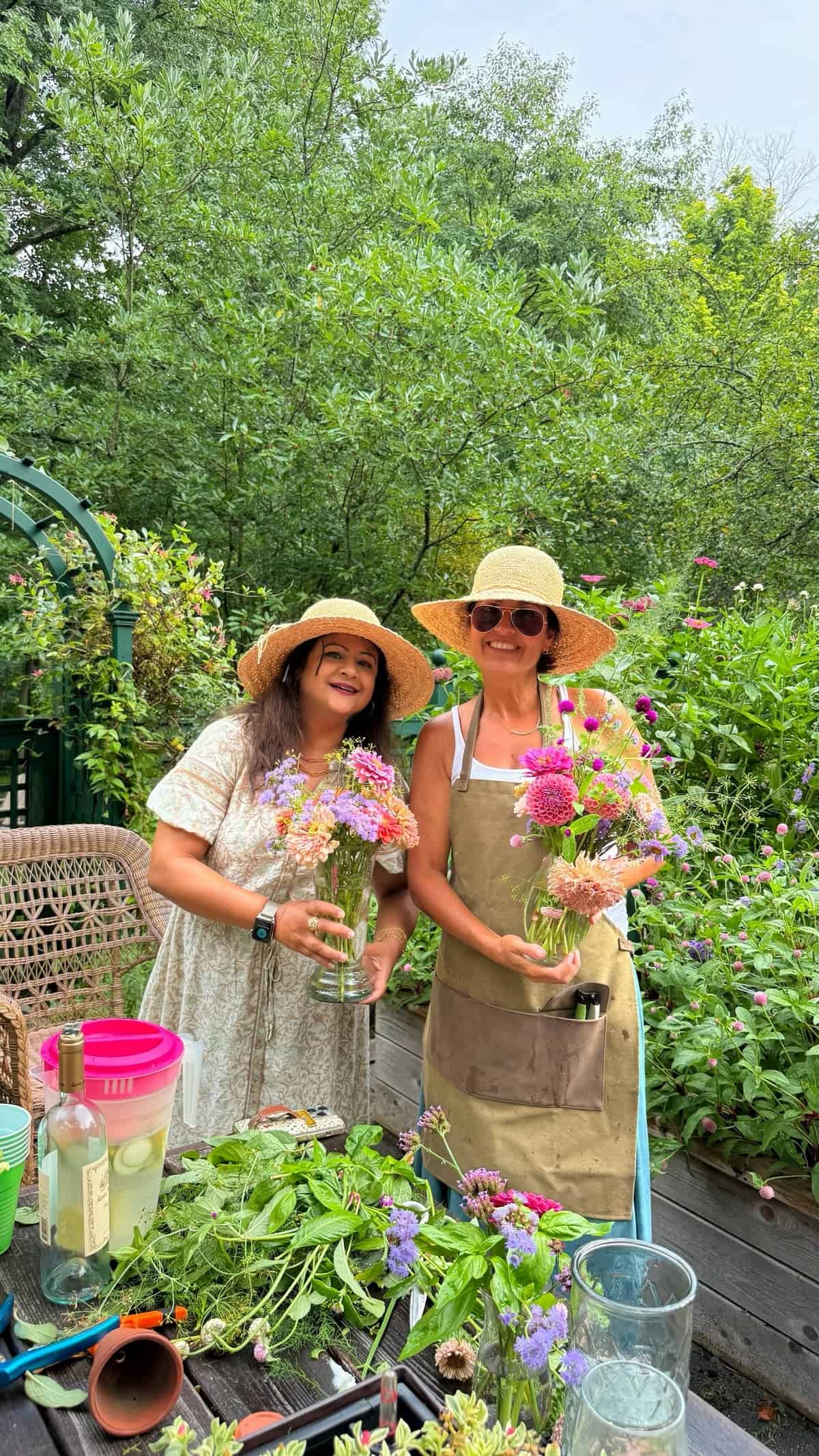 Two women in sun hats stand outdoors in a lush garden, smiling while holding colorful flower arrangements. A table in front of them displays flowers, wine, and garden tools.