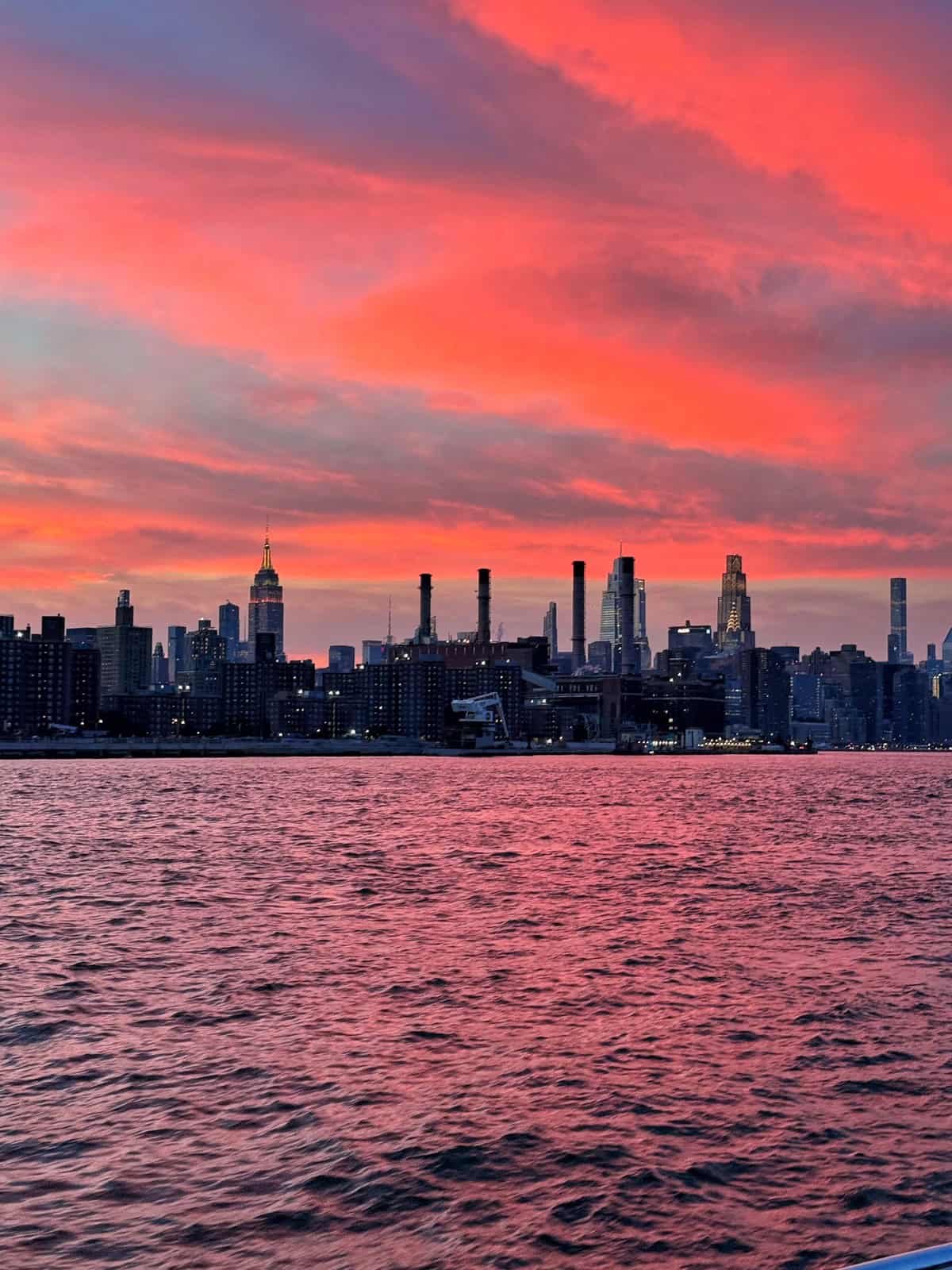 The New York City skyline is pictured at sunset, with vivid pink and orange clouds above and the city’s buildings silhouetted against the colorful sky, reflected in the water below.