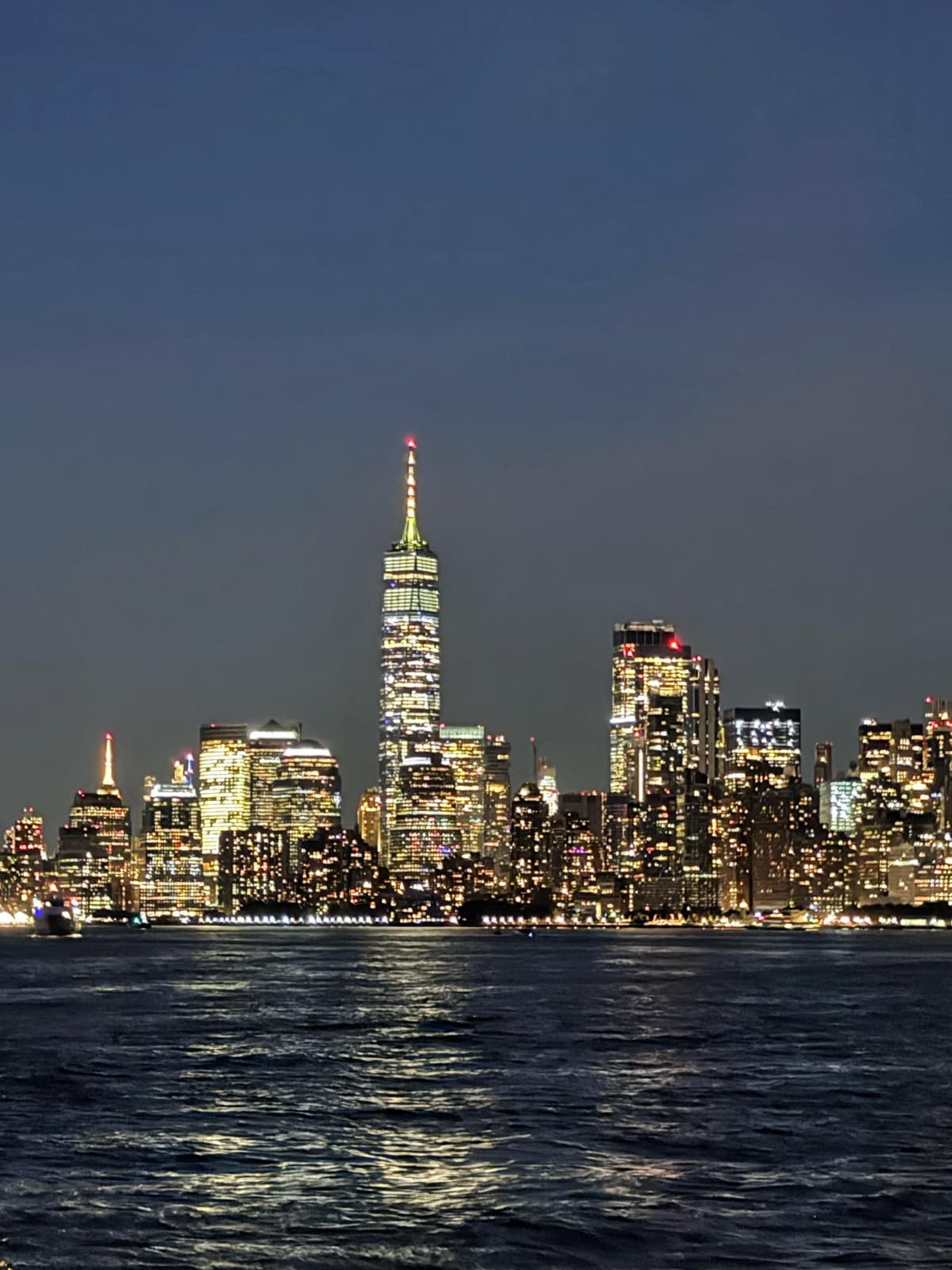 Nighttime cityscape with illuminated skyscrapers reflecting on the water, featuring a tall building in the center with a lit spire against a dark, clear sky.