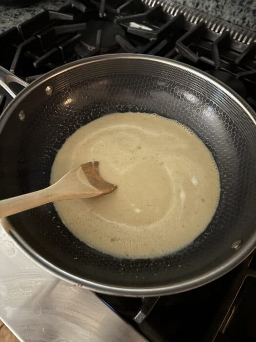 A wooden spoon stirs a light-colored sauce or roux in a nonstick pan on a stovetop. The burner grates and stove controls are visible in the background.