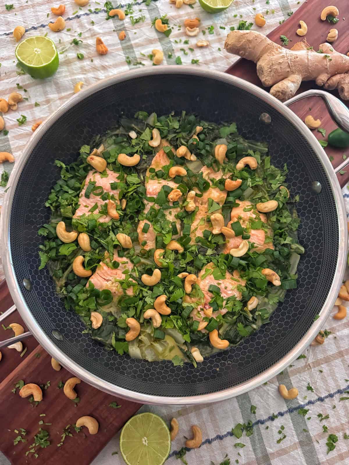 Salmon fillets in a pan topped with chopped herbs and cashew nuts, surrounded by lime halves, ginger roots, and scattered cashews on a patterned tablecloth.