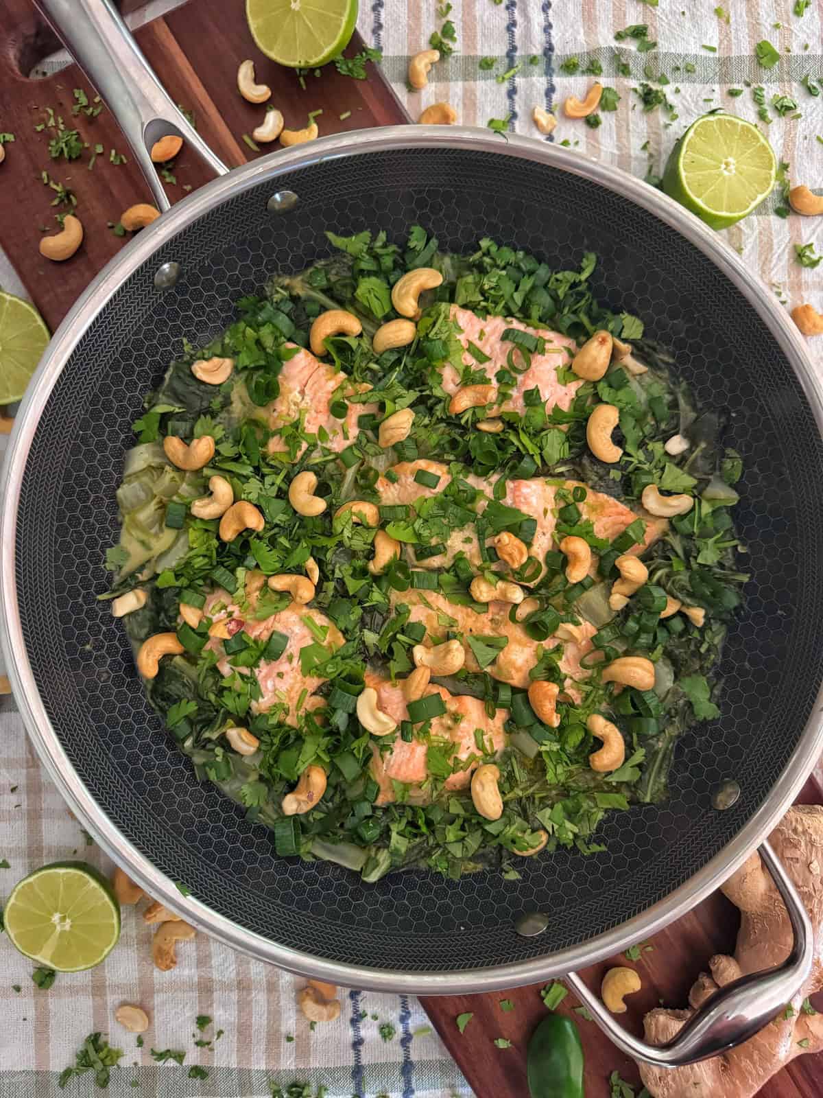 Salmon fillets in a pan topped with fresh herbs and cashews, surrounded by lime halves and scattered greens on a patterned tablecloth.