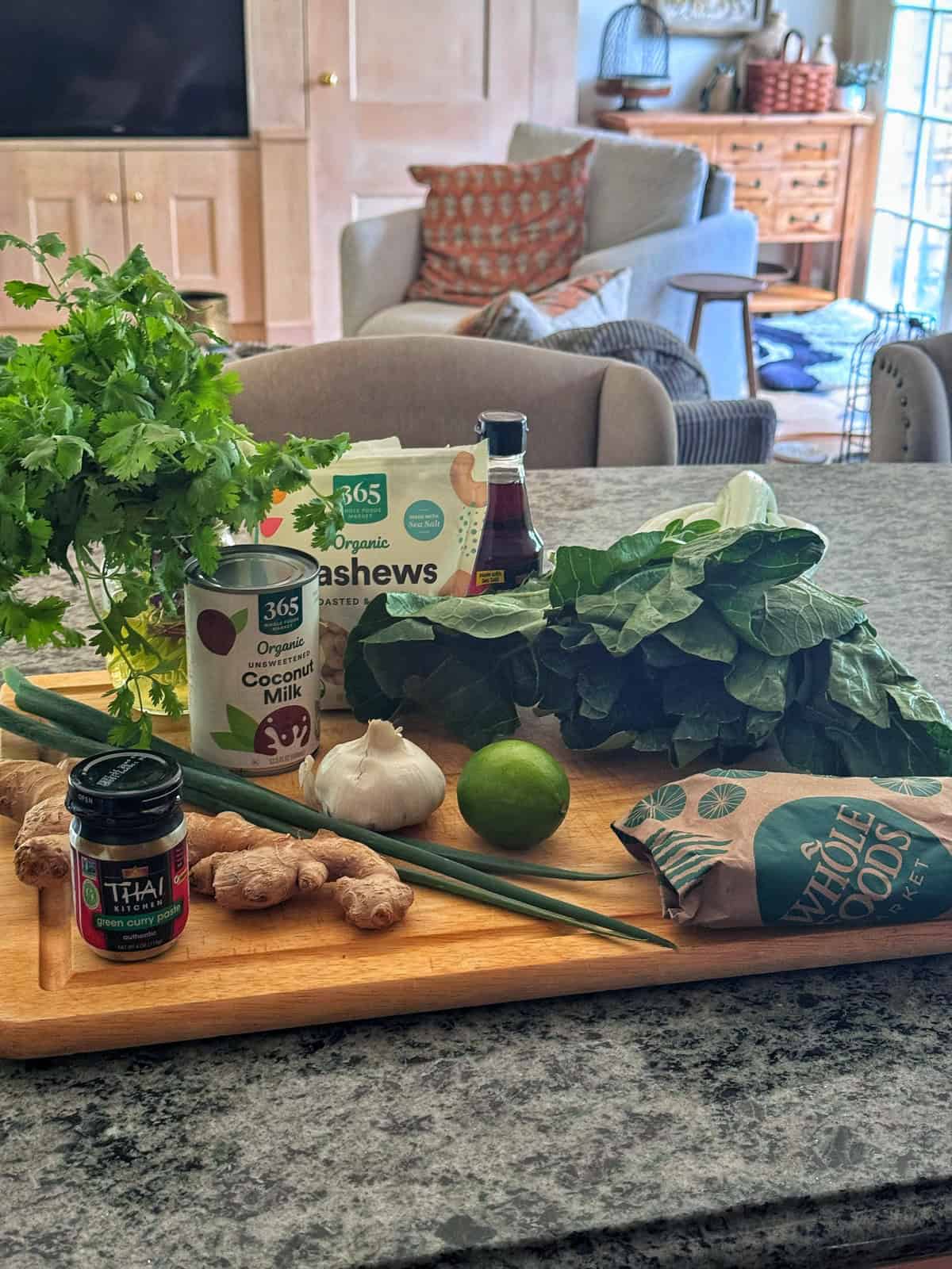 A wooden board on a kitchen counter holds fresh herbs, spinach, garlic, ginger, a lime, green onions, coconut milk, cashews, soy sauce, and Thai red curry paste. A living room is seen in the background.