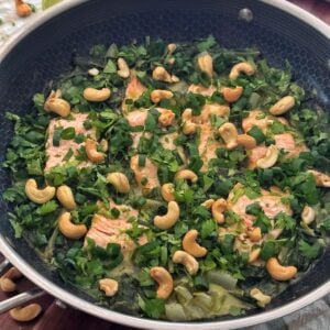 Salmon fillets in a skillet, topped with fresh herbs and cashews, with lime halves and a ginger root nearby on a wooden surface.