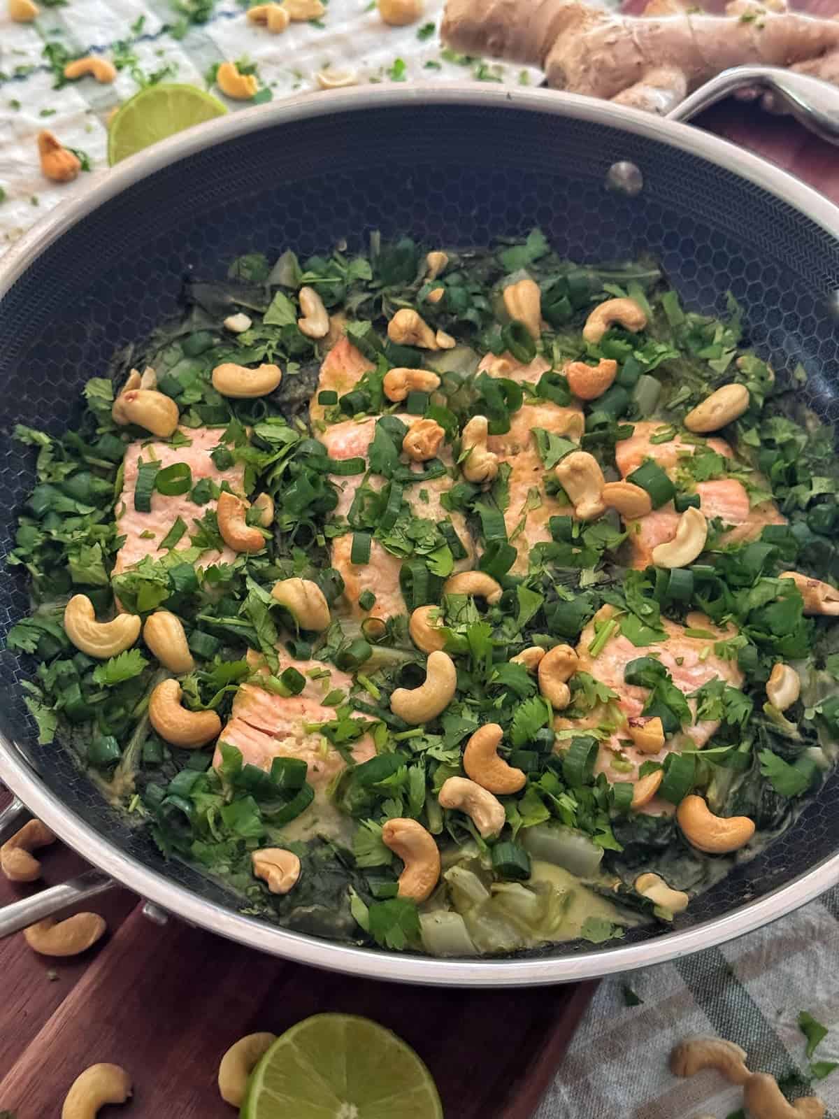 Salmon fillets in a skillet, topped with fresh herbs and cashews, with lime halves and a ginger root nearby on a wooden surface.