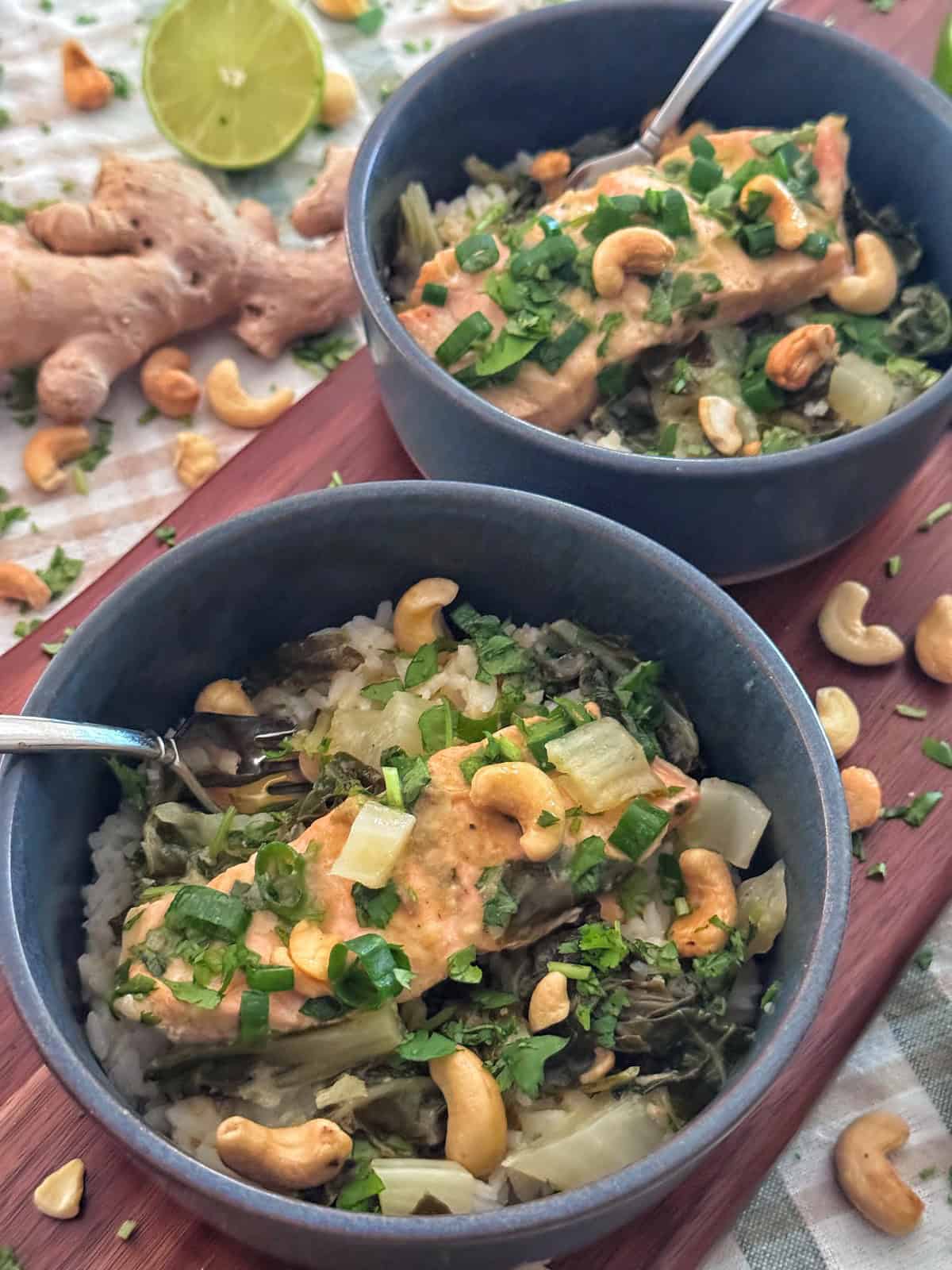 Two bowls of rice topped with vegetables, a creamy sauce, chopped herbs, and cashews. A fork rests in each bowl. Garnishes and ingredients like cashews, ginger, and lime are scattered on the table nearby.