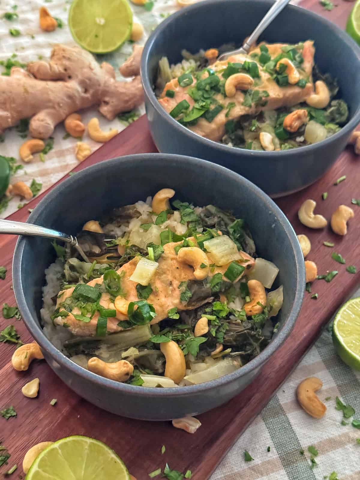 Two bowls of rice topped with vegetables, greens, creamy sauce, cashews, and chopped herbs. Slices of lime, ginger, whole cashews, and herbs are scattered on the wooden surface around the bowls.