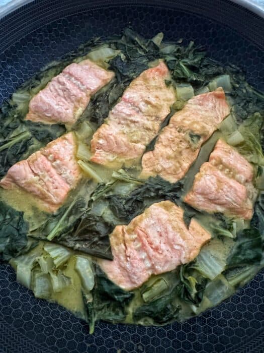 Six pieces of cooked salmon are arranged in a pan with leafy greens and a light, creamy sauce.
