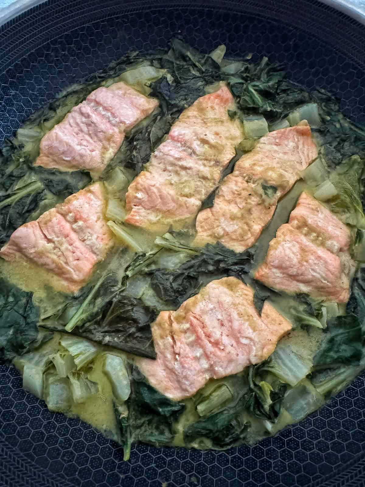 Six pieces of cooked salmon are arranged in a pan with leafy greens and a light, creamy sauce.
