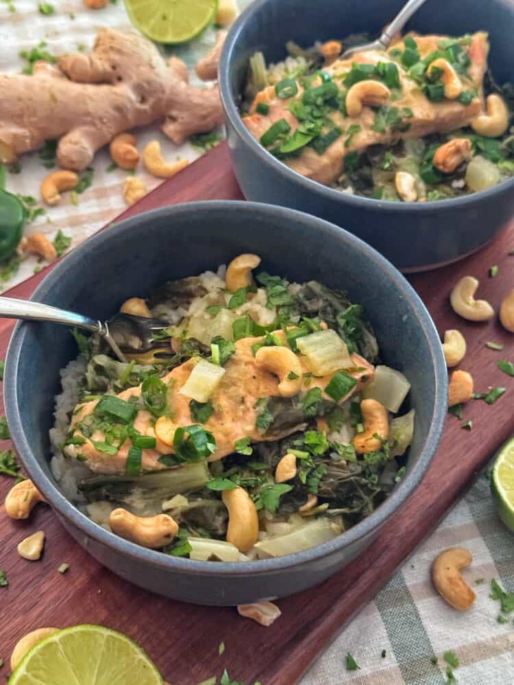 Two bowls with salmon and bok choy with green curry sauce that are filled with rice topped with green vegetables, baked fish, chopped herbs, and cashews, placed on a wooden tray. Lime slices, cashews, and ginger are scattered around on a checkered cloth.