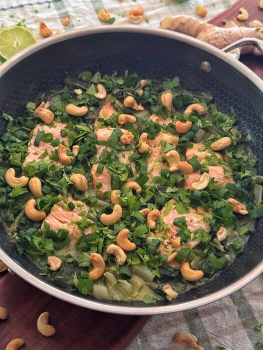 Salmon fillets in a pan topped with fresh herbs and cashews, surrounded by leafy greens and garnished with chopped green onions, on a checkered tablecloth.