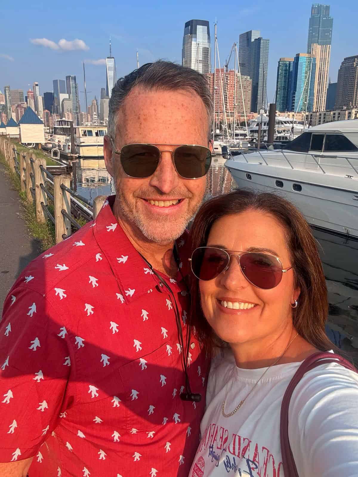 A smiling man christopher ling and woman stacy ling in sunglasses pose for a selfie at a marina with boats and tall city buildings in the background, under a clear blue sky.