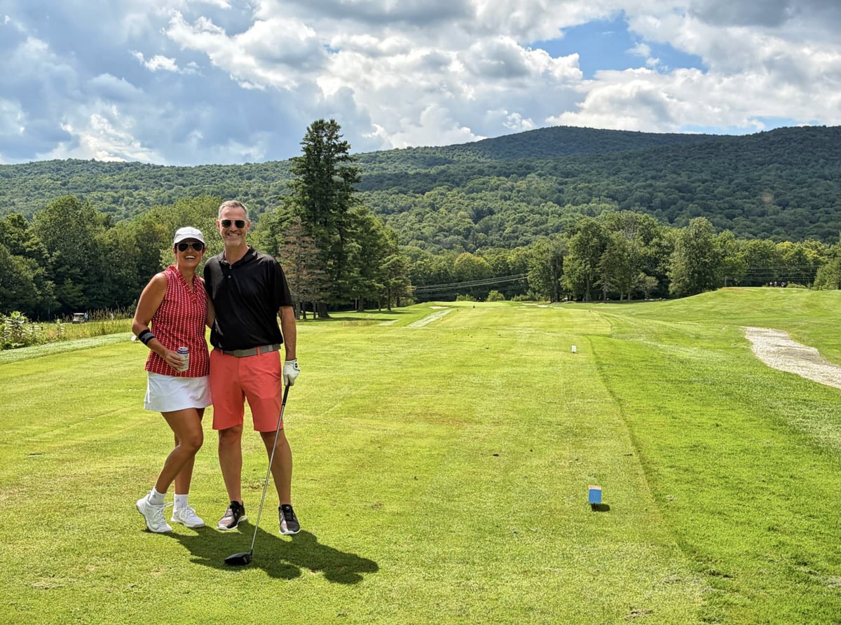 Two people smiling on a golf course, holding golf clubs. Green grass, trees, and hills stretch behind them under a partly cloudy sky.