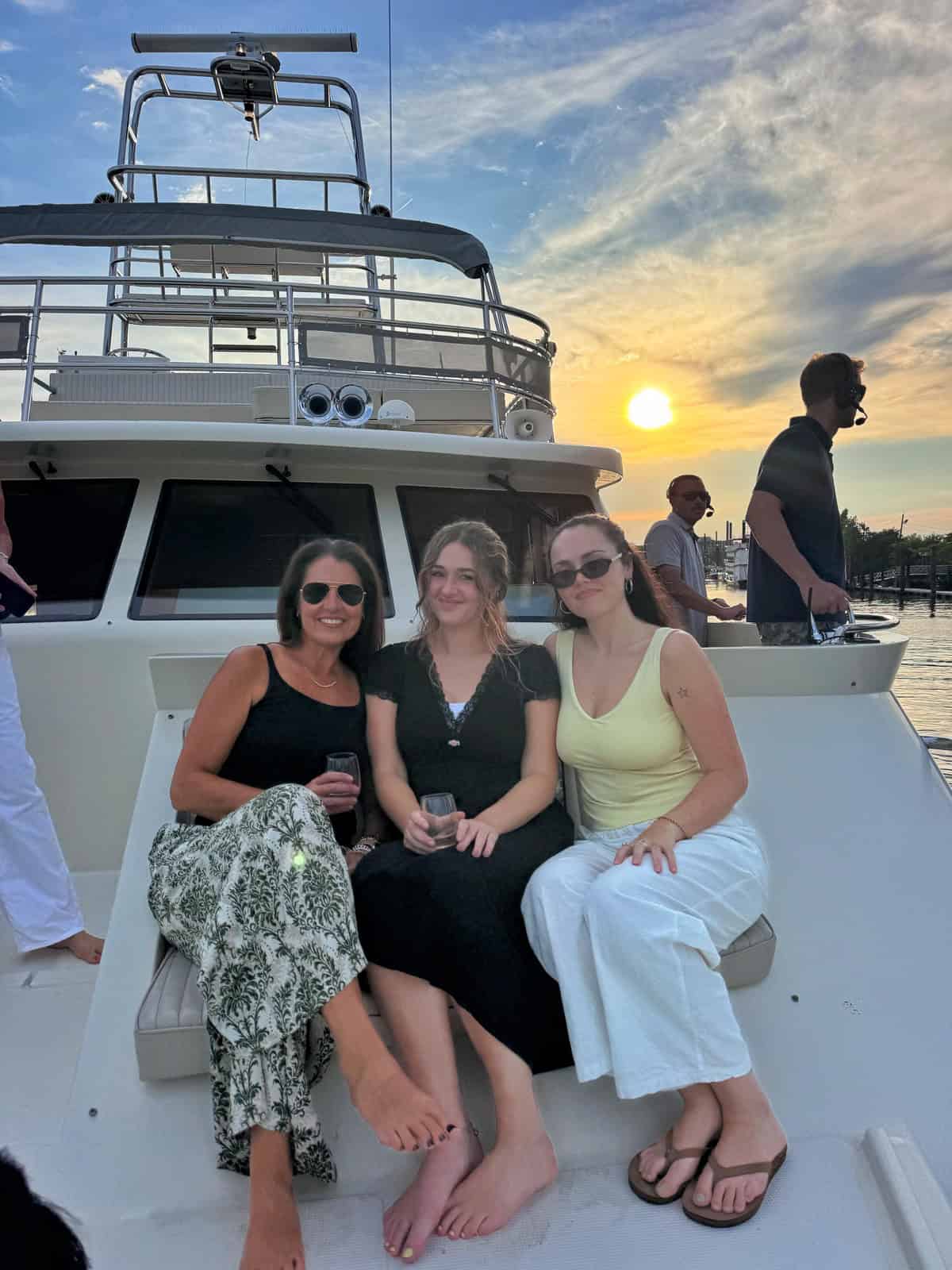 Three women sit barefoot on a boat at sunset, smiling and holding drinks. Other people stand in the background, and the sky is partly cloudy with the sun low on the horizon.