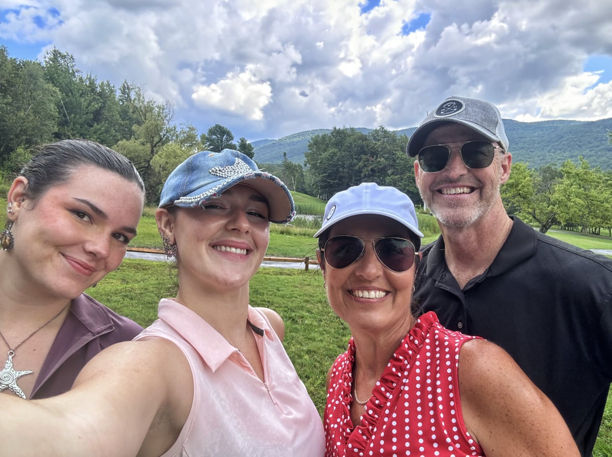 Four smiling adults pose for a selfie outdoors on a green lawn, with trees and mountains in the background under a partly cloudy sky.