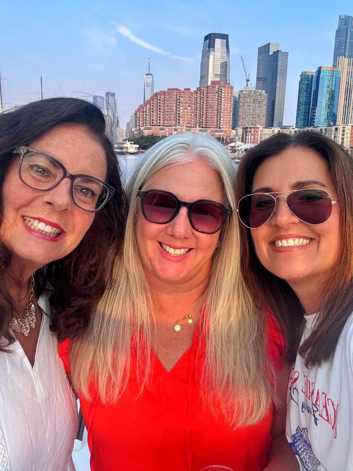 Three smiling women wearing sunglasses pose for a selfie outdoors with tall city buildings and a blue sky in the background.