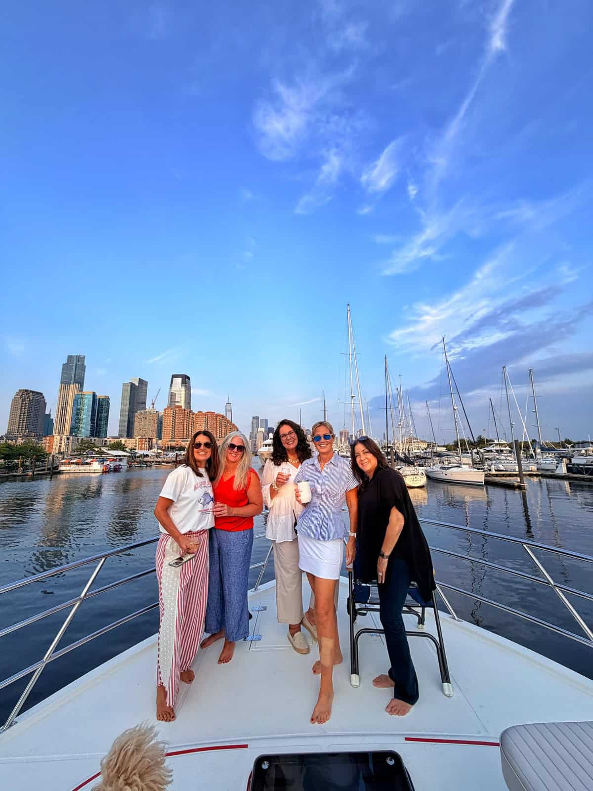 Five women smile and pose together on the bow of a boat, with a marina, sailboats, and city skyline in the background under a bright blue sky.