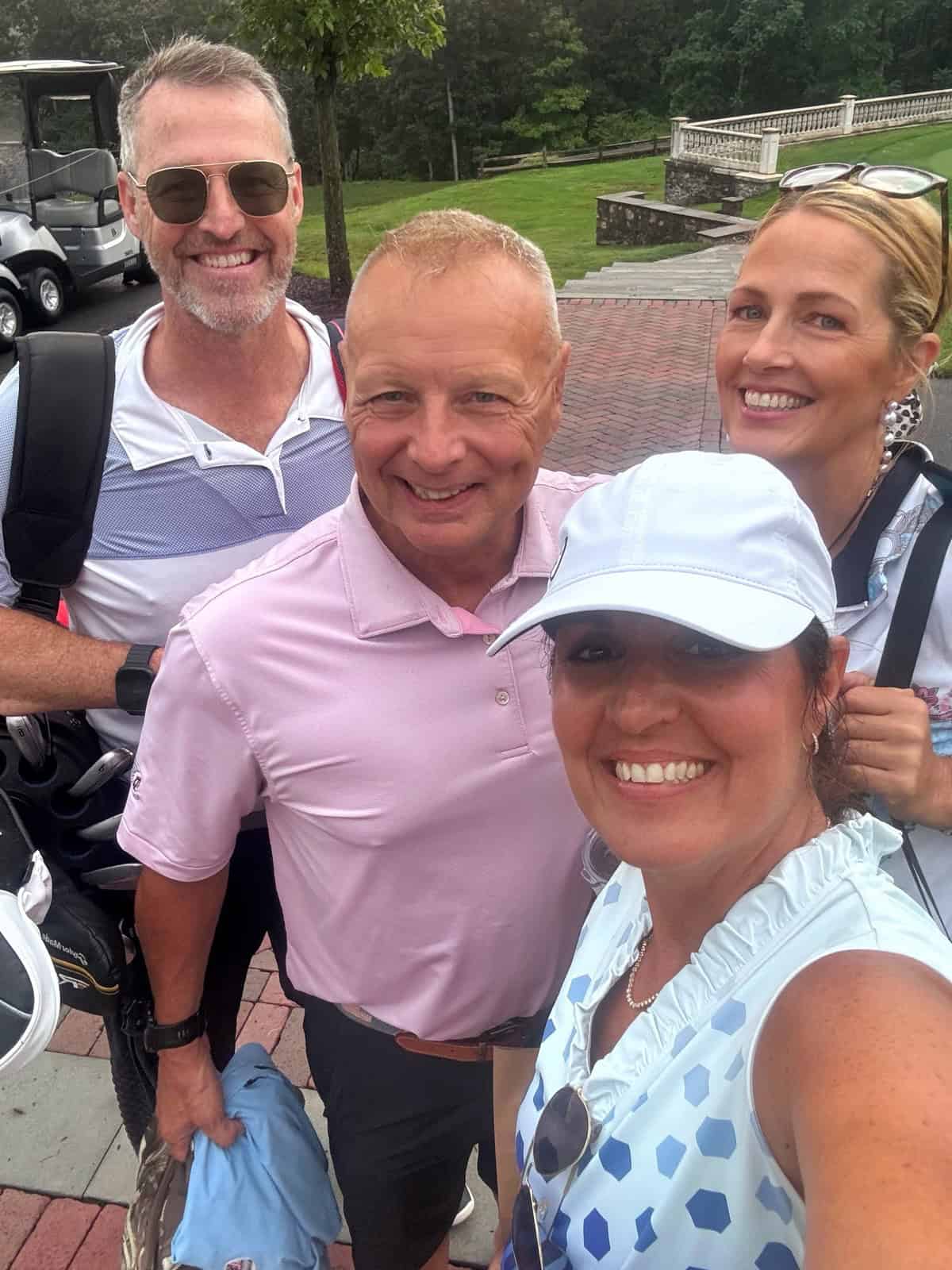 Four adults smile for a selfie outdoors on a brick path, with golf carts and green grass in the background. Two women are in the foreground; two men are behind them. All are dressed in golf attire.