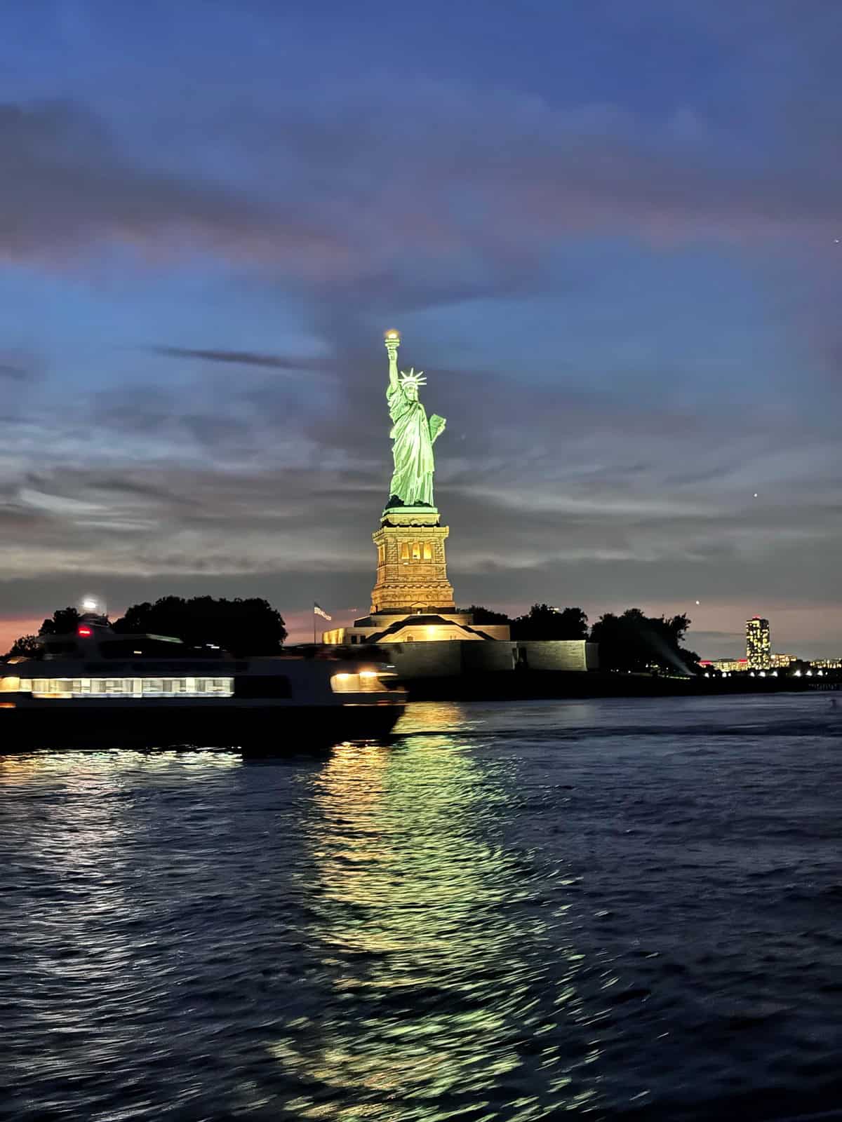 The Statue of Liberty illuminated at dusk, standing on Liberty Island with a boat passing by and water reflecting the statue’s light.