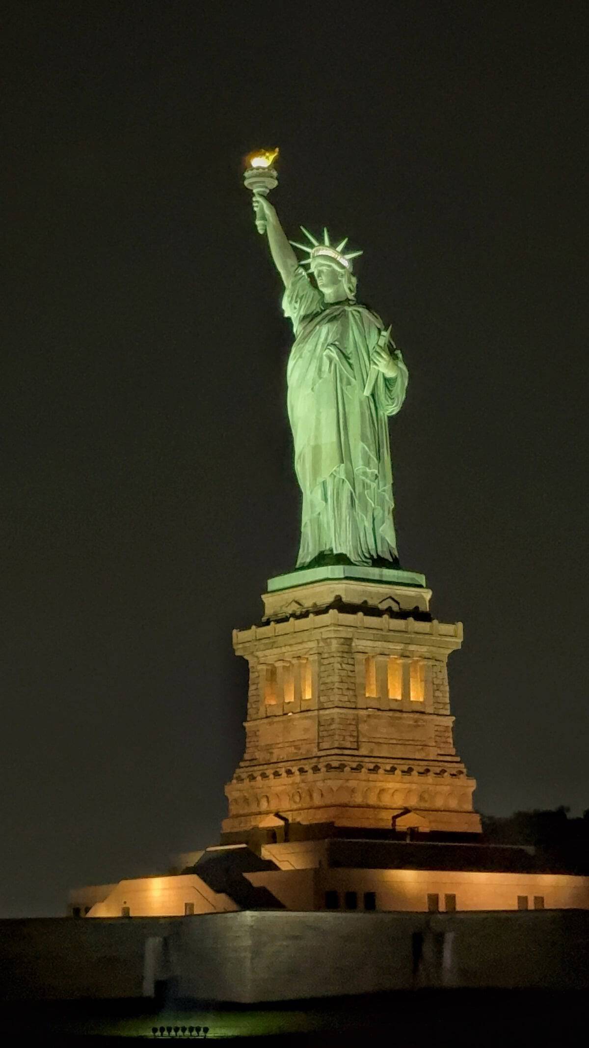The Statue of Liberty stands illuminated at night, with its torch raised high and the pedestal lit by golden lights against a dark sky.