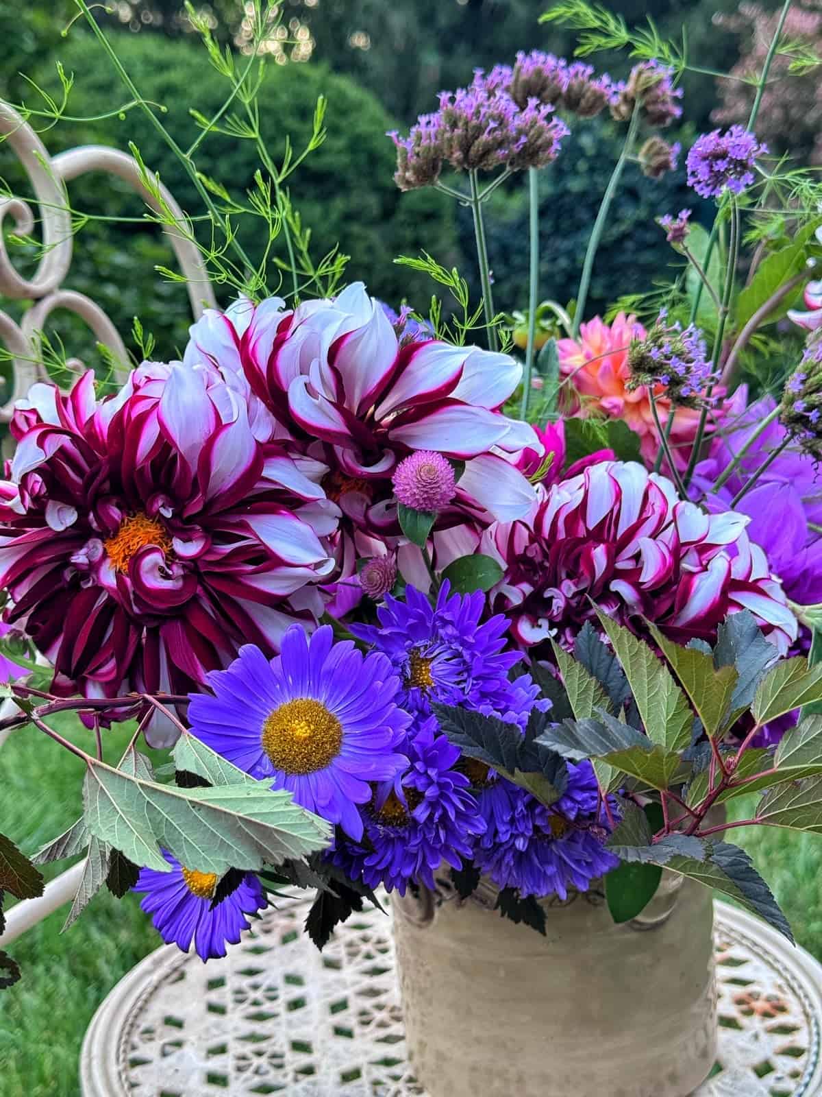 A close-up of a floral arrangement with purple and white dahlias, purple asters, and greenery in a cream-colored vase on an outdoor table, with a blurred garden background.