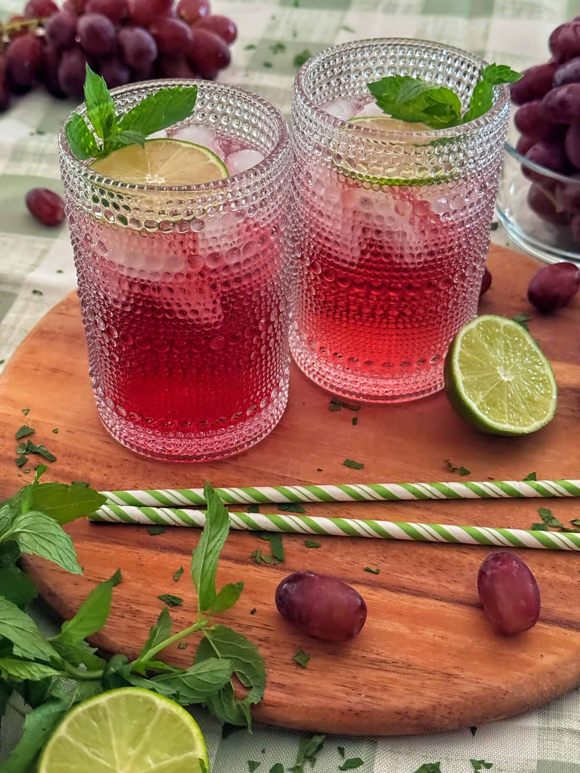 Two textured glasses with a classic transfusion cocktail recipe, garnished with lime slices and mint, sit on a wooden board. Grapes, lime halves, mint leaves, and striped straws are arranged around them on a checkered tablecloth.