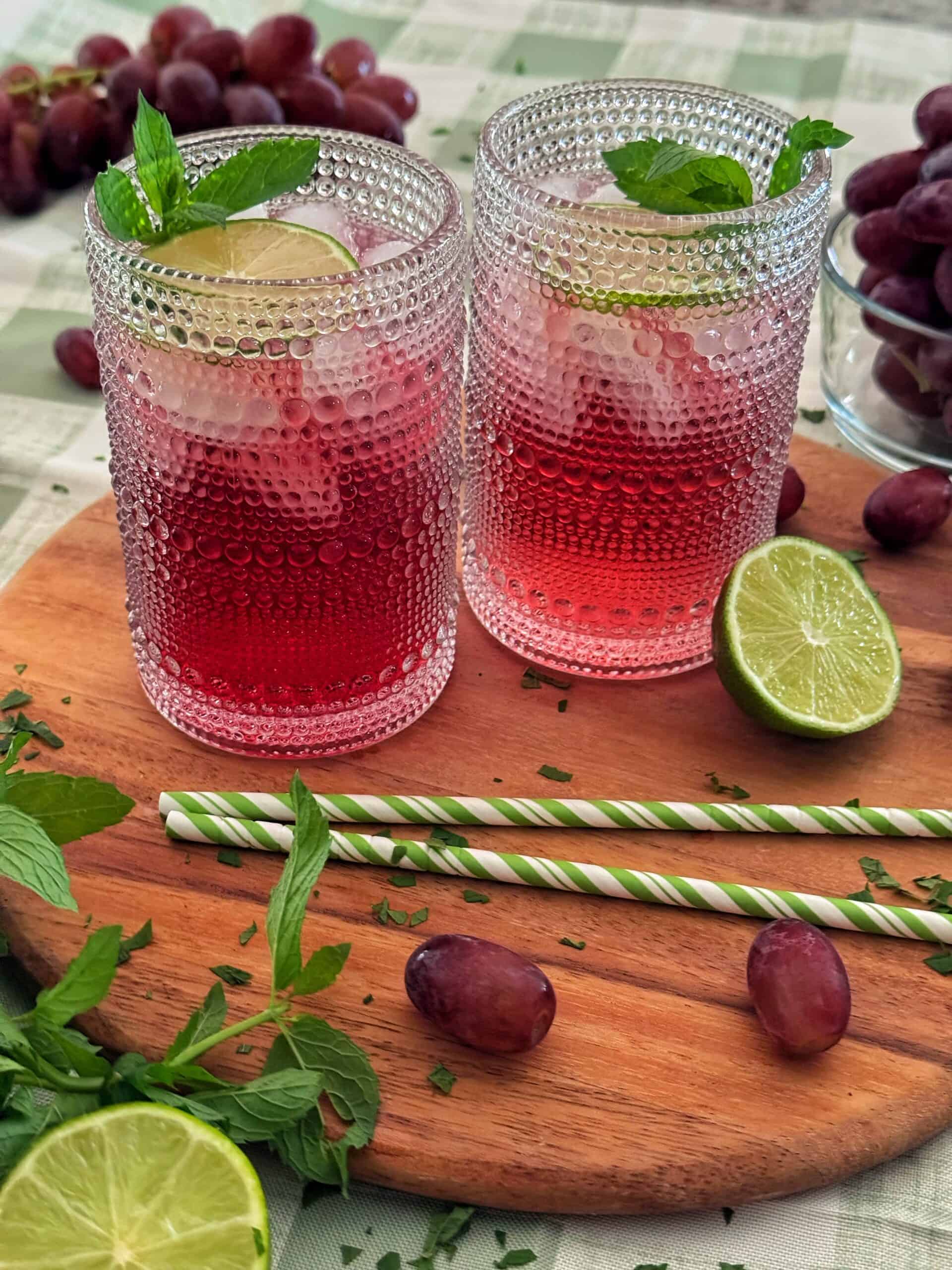 Two textured glasses of a classic transfusion cocktail with ice, garnished with lime slices and mint, sit on a wooden tray. Fresh grapes, lime halves, mint leaves, and striped paper straws are nearby on a patterned tablecloth.