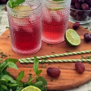 Two glasses of a classic transfusion cocktail garnished with lime and mint, placed on a wooden board with grapes, lime halves, mint leaves, and striped straws, with a bowl of grapes in the background.