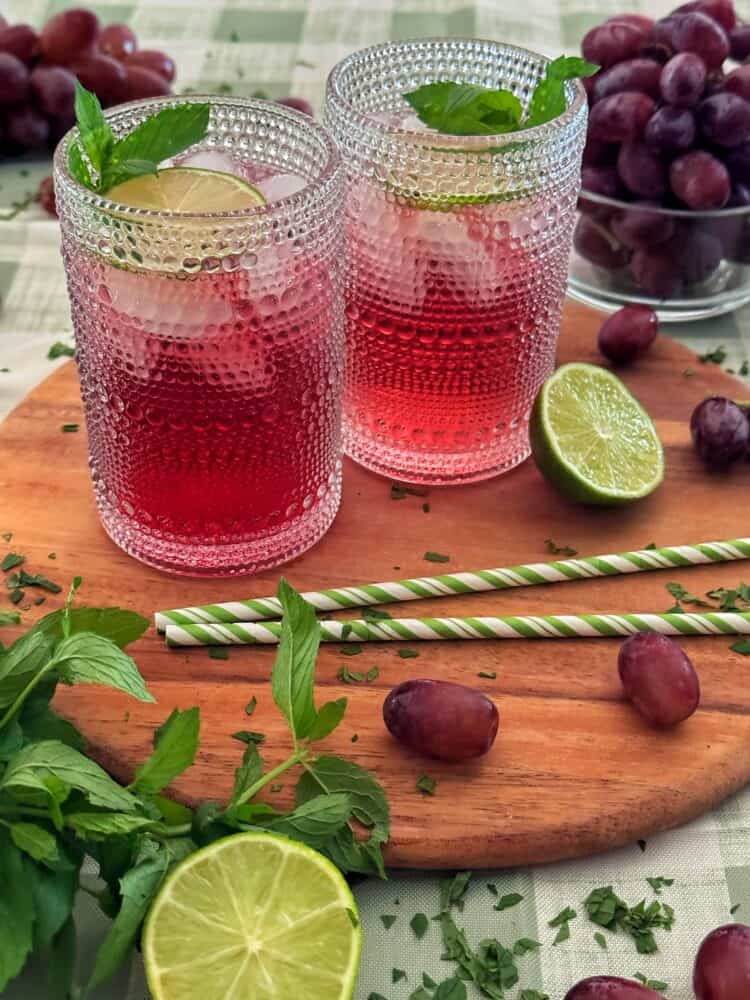 Two glasses of a classic transfusion cocktail garnished with lime and mint, placed on a wooden board with grapes, lime halves, mint leaves, and striped straws, with a bowl of grapes in the background.