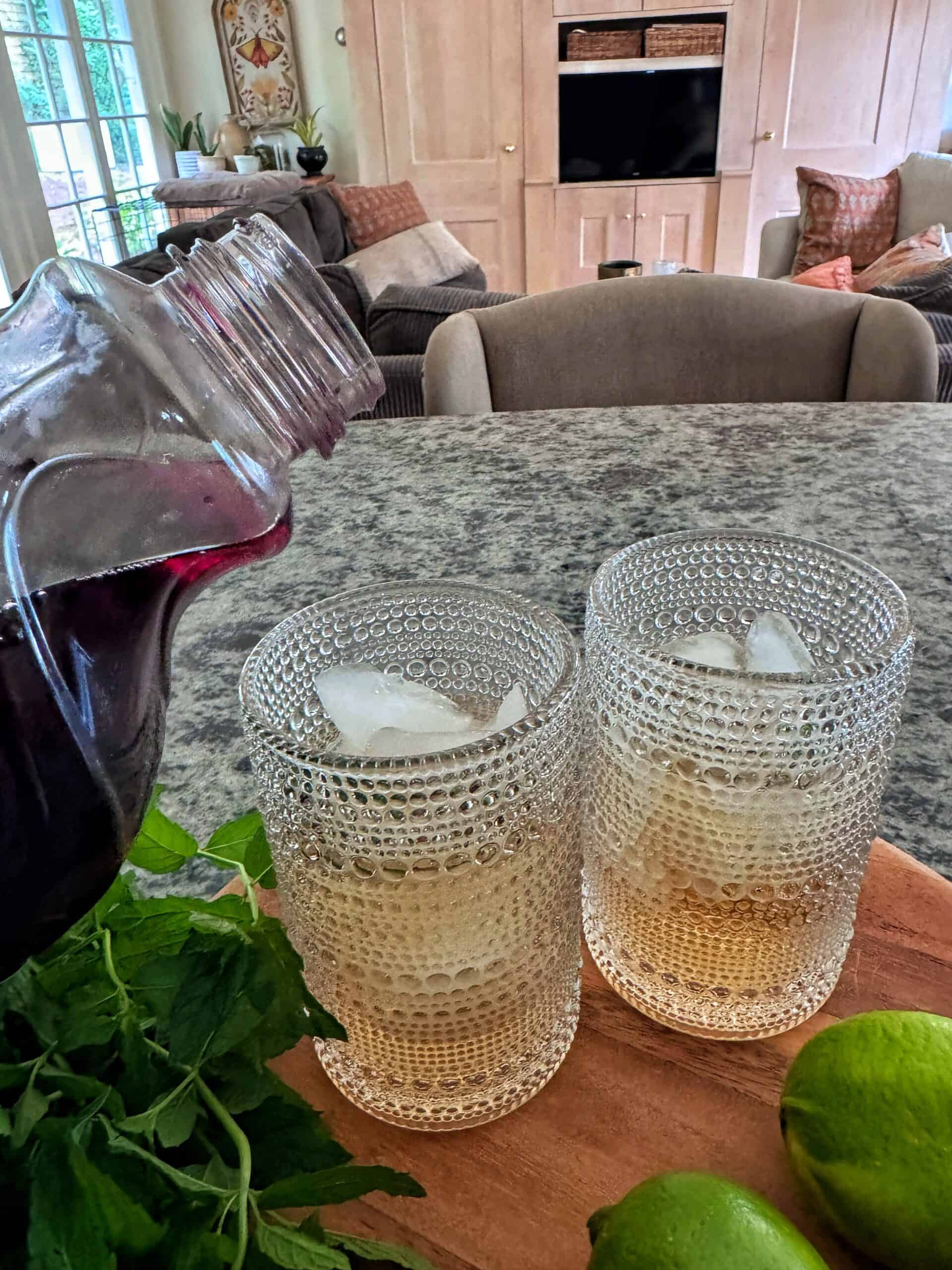 A hand pours a dark liquid from a bottle into one of two glasses with ice on a countertop. Fresh mint and limes are nearby. A cozy living room is visible in the background.