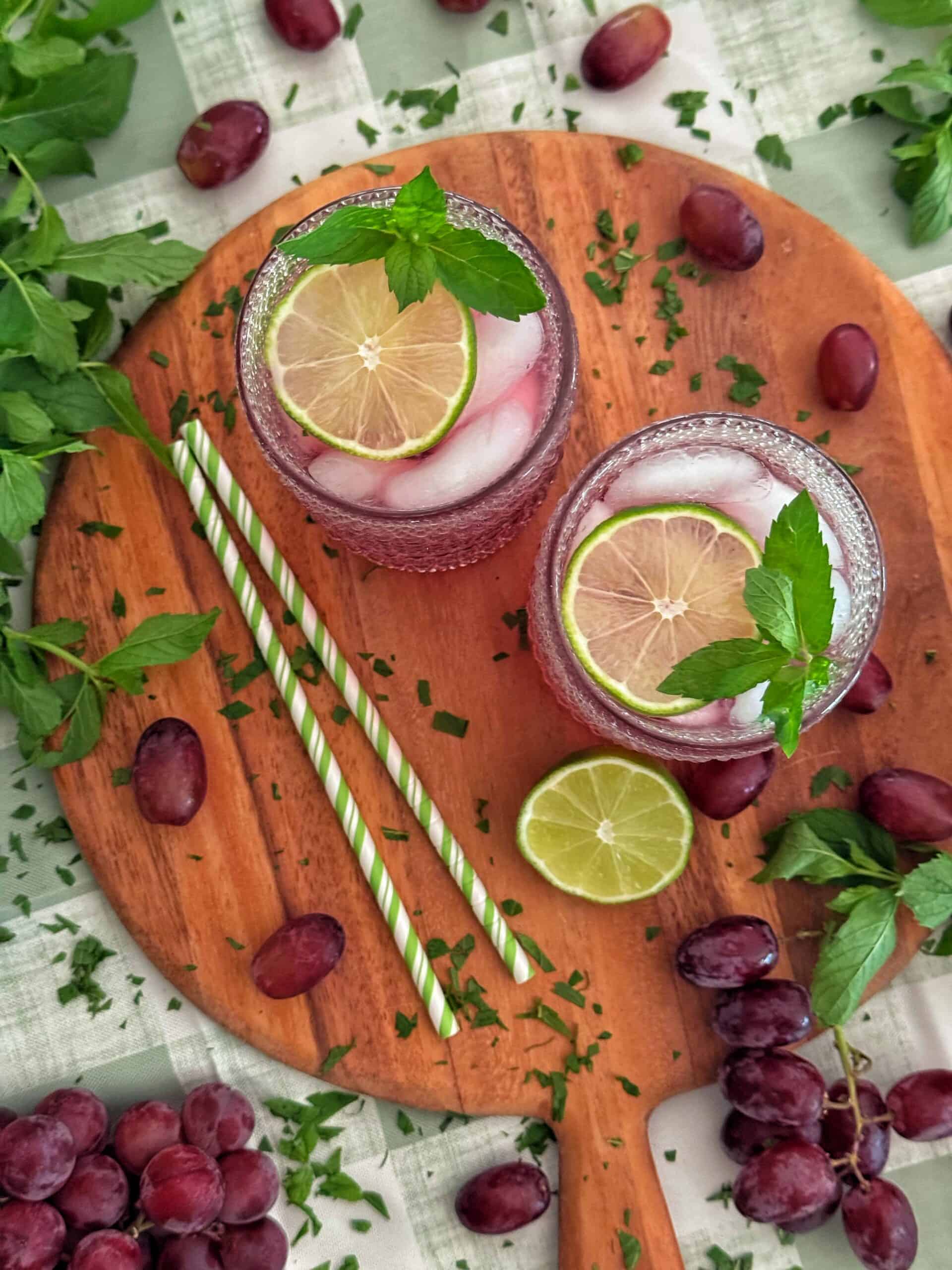 Two glasses of iced grape drink with lime slices and mint on a wooden board, surrounded by grapes, lime, herbs, and striped straws.