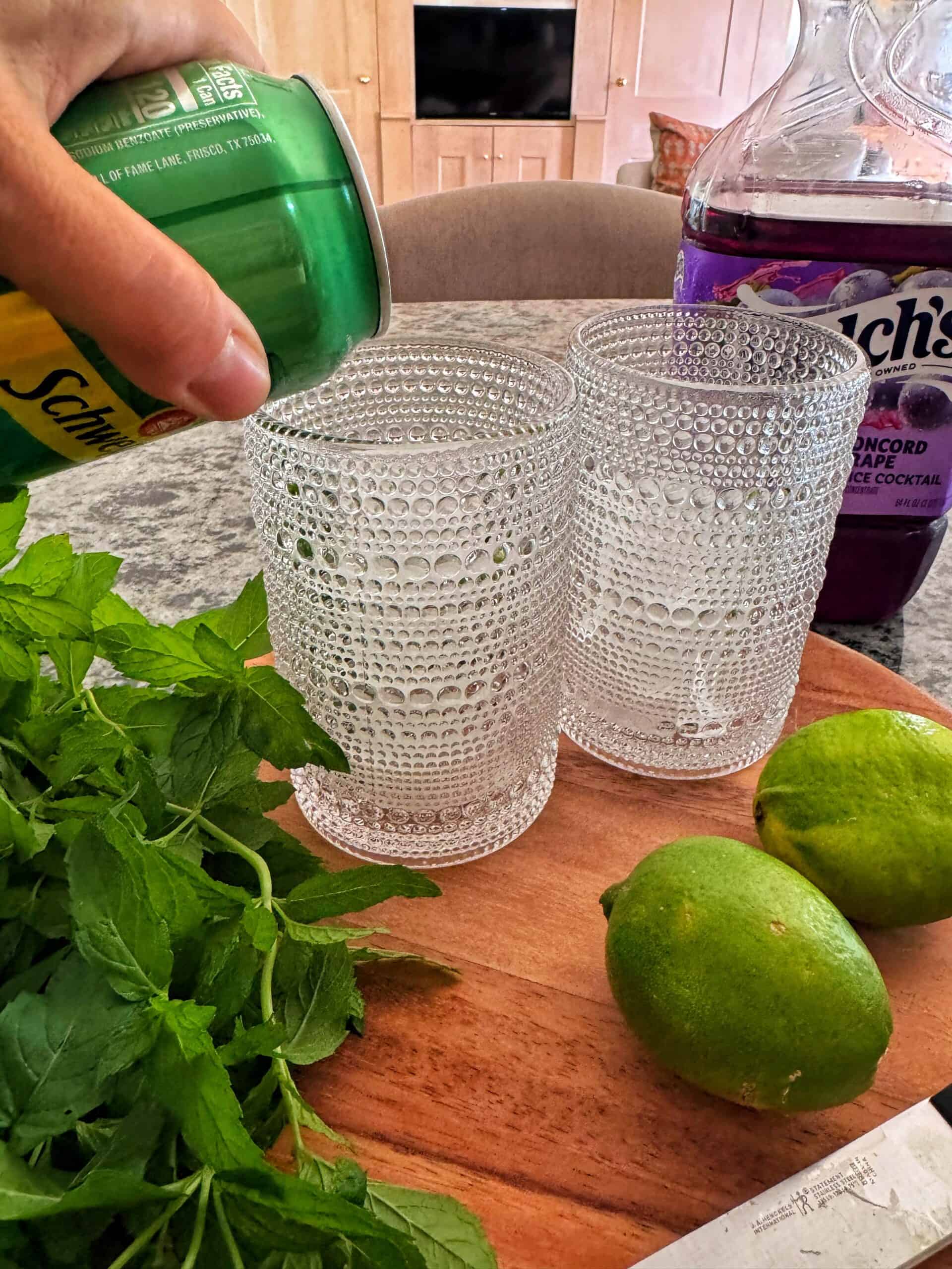 A hand pours soda from a green can into textured glasses on a wooden tray with mint, two limes, and a purple bottle of Welch’s grape juice nearby. A kitchen background is visible.