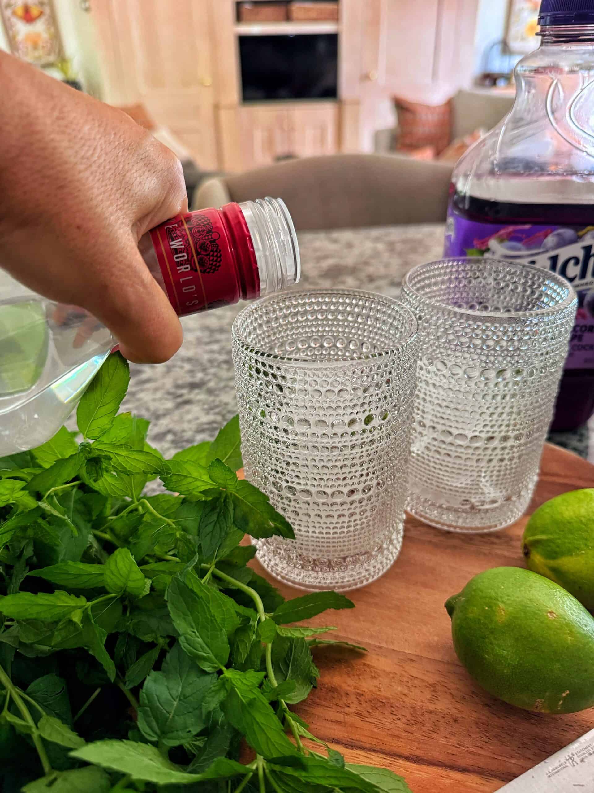 A hand pours sparkling water into a textured glass. Next to it are another glass, fresh mint, limes, and a bottle of grape juice on a kitchen counter.