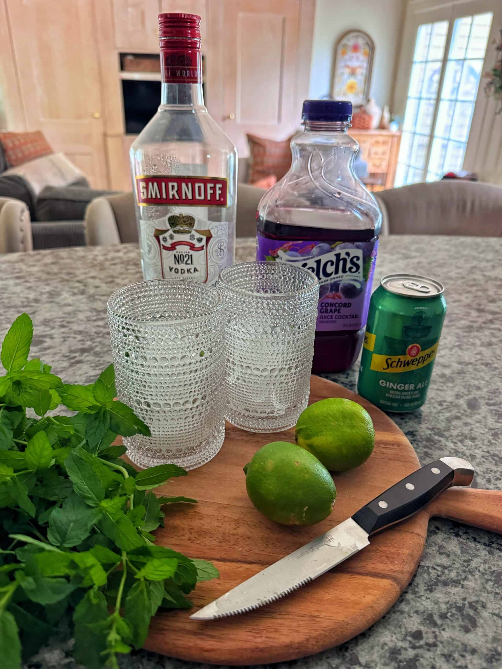 A kitchen counter displays vodka, grape juice, ginger ale, mint, limes, a knife, and two textured glasses, suggesting cocktail preparation.