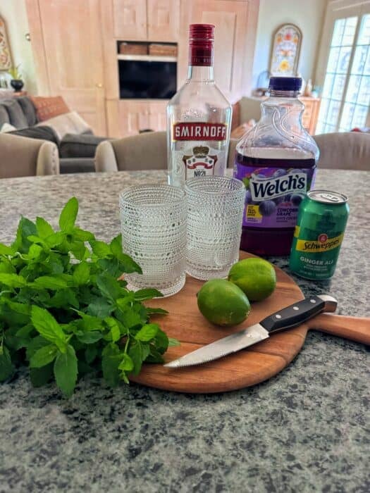 A cutting board with mint, two limes, and a knife sits on a counter next to two glasses, a bottle of Smirnoff vodka, Welch’s grape juice, and a can of Schweppes ginger ale.