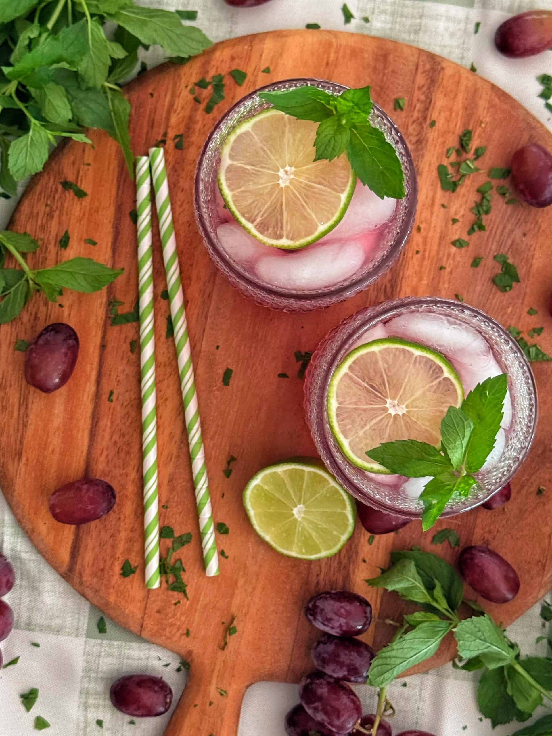 Two glasses of the classic transfusion cocktail topped with lime slices and mint sit on a wooden board, surrounded by grapes, mint leaves, and two striped straws.