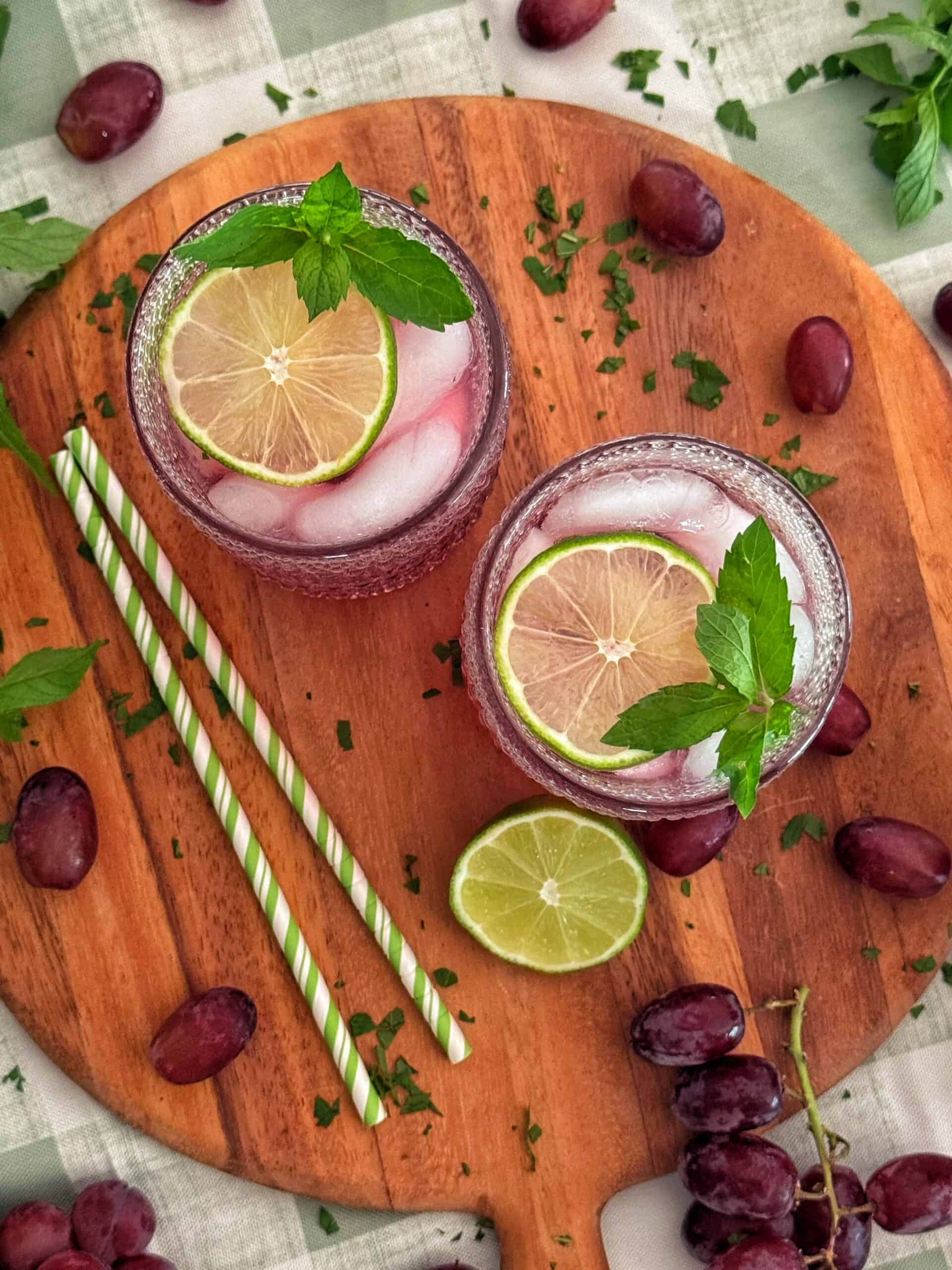 Classic transfusion cocktail in two glasses with ice, lime slices, and mint, sitting on a wooden board with grapes, lime, and green-striped straws scattered around. A bunch of grapes and herbs are also visible.