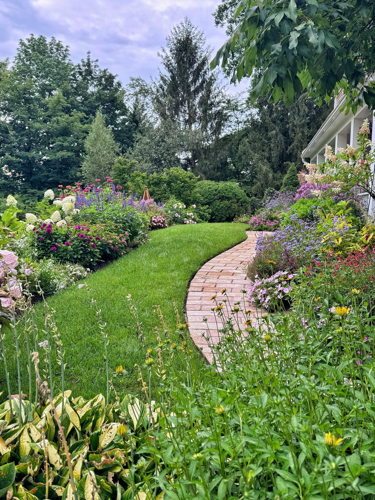 A curved brick path borders a lush green lawn, surrounded by vibrant flowering plants and tall trees in a garden next to a house.