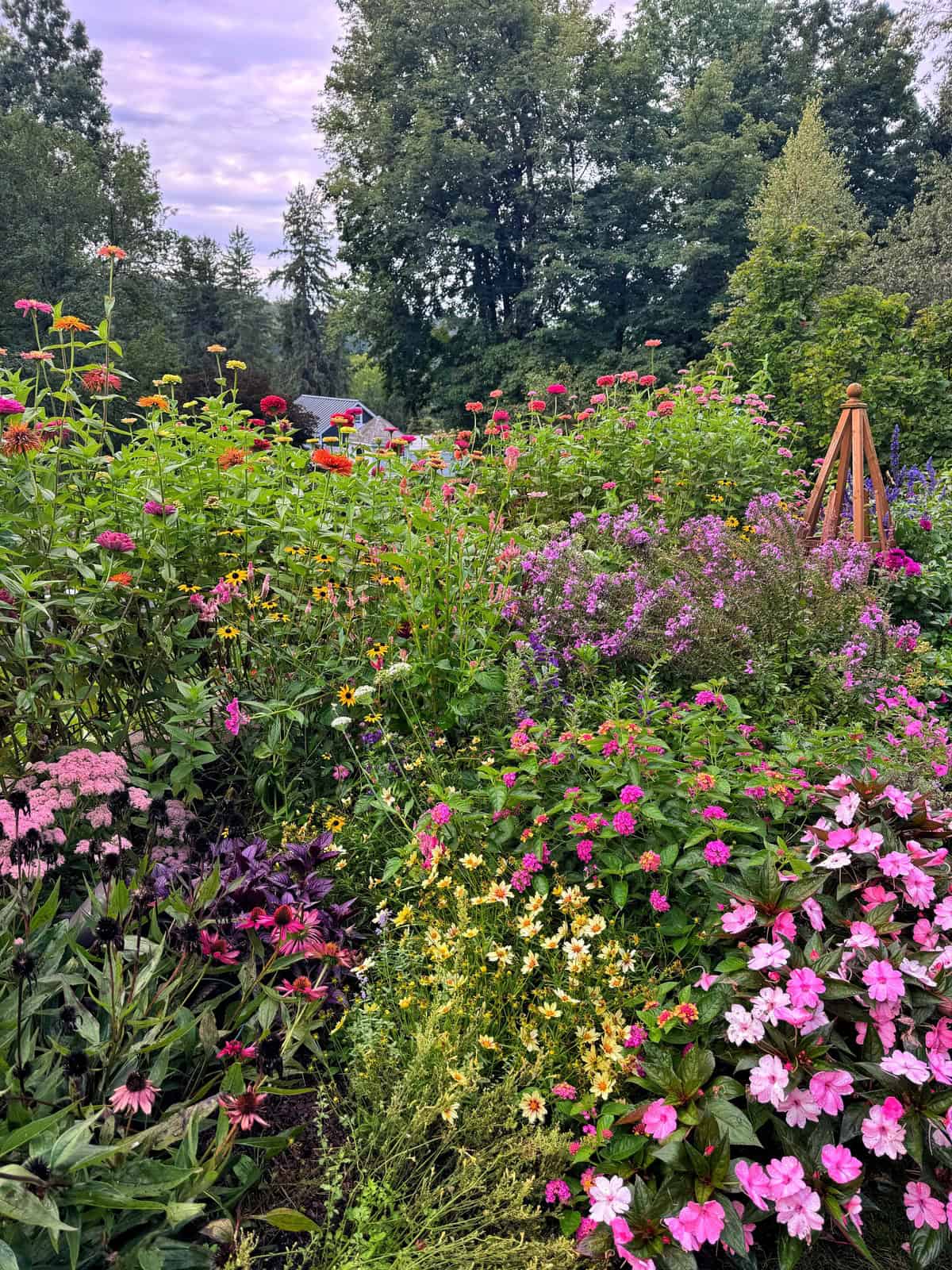 A lush garden is filled with colorful blooming flowers in pink, yellow, purple, and red, surrounded by green trees, under a partly cloudy sky. A wooden garden obelisk stands among the flowers.