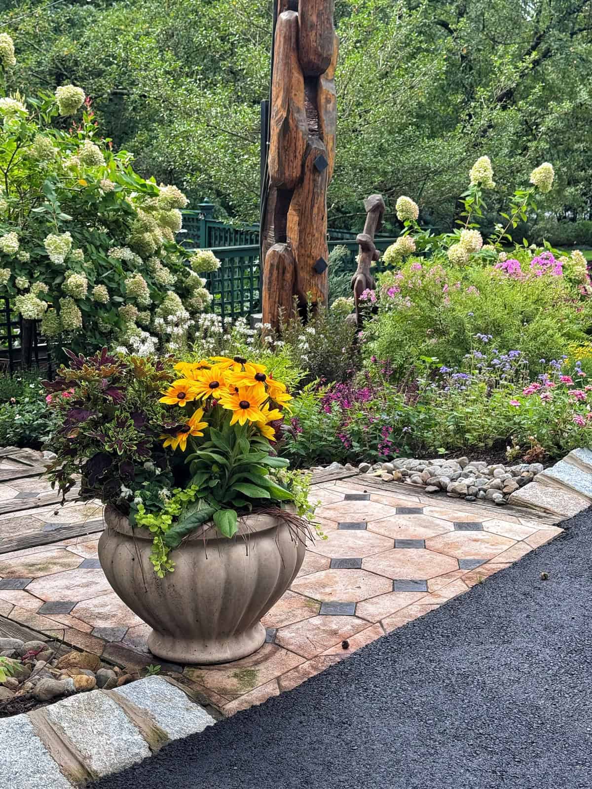 Large stone planter with bright yellow flowers sits on a patterned patio, surrounded by blooming garden beds, green shrubs, and carved wooden sculpture in the background.