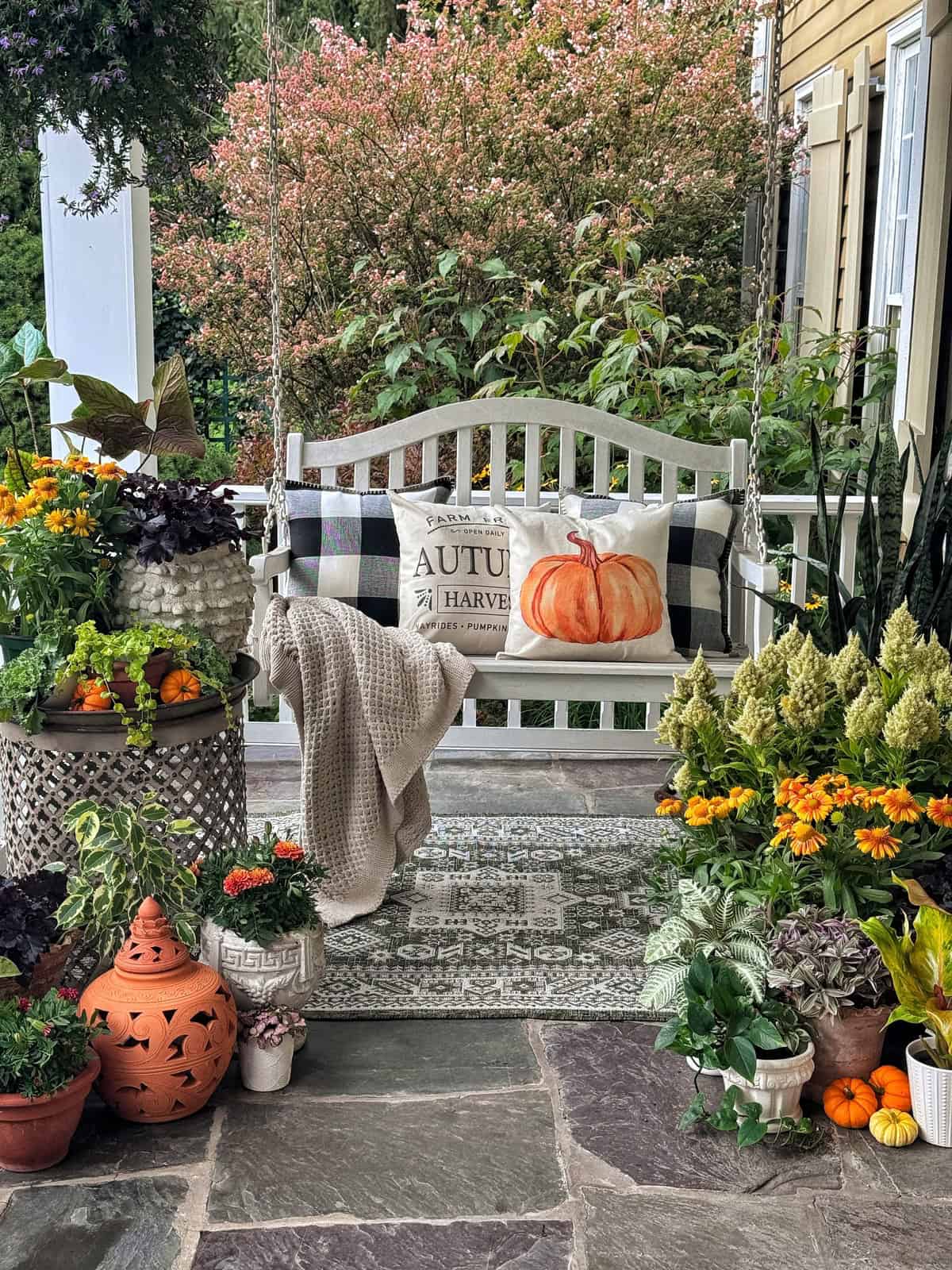 A cozy porch swing with autumn-themed pillows and blanket, surrounded by vibrant potted flowers and greenery. The scene has a welcoming, fall harvest vibe with pumpkins and a patterned outdoor rug on a stone floor.