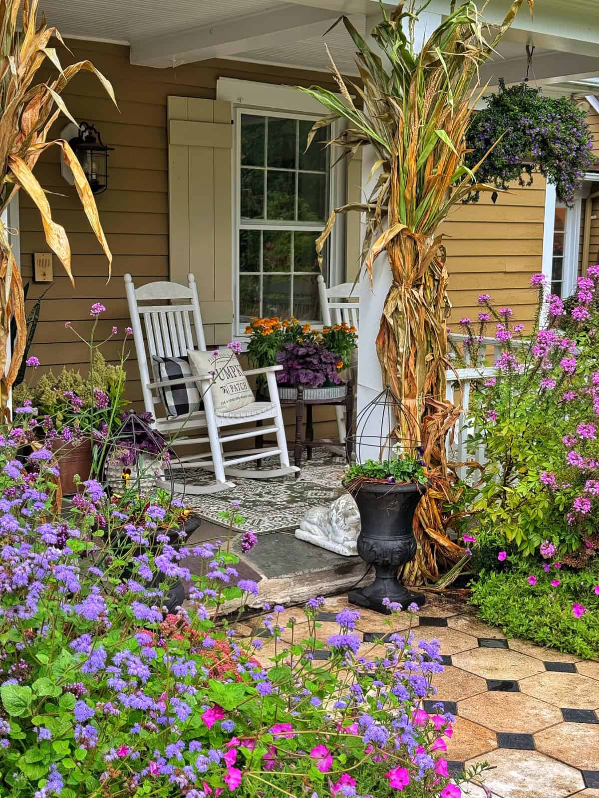 A cozy porch with white rocking chairs, potted flowers, dried cornstalks, and a stone path surrounded by vibrant purple and pink flowers. A welcome sign is on one chair, creating a warm, inviting atmosphere.