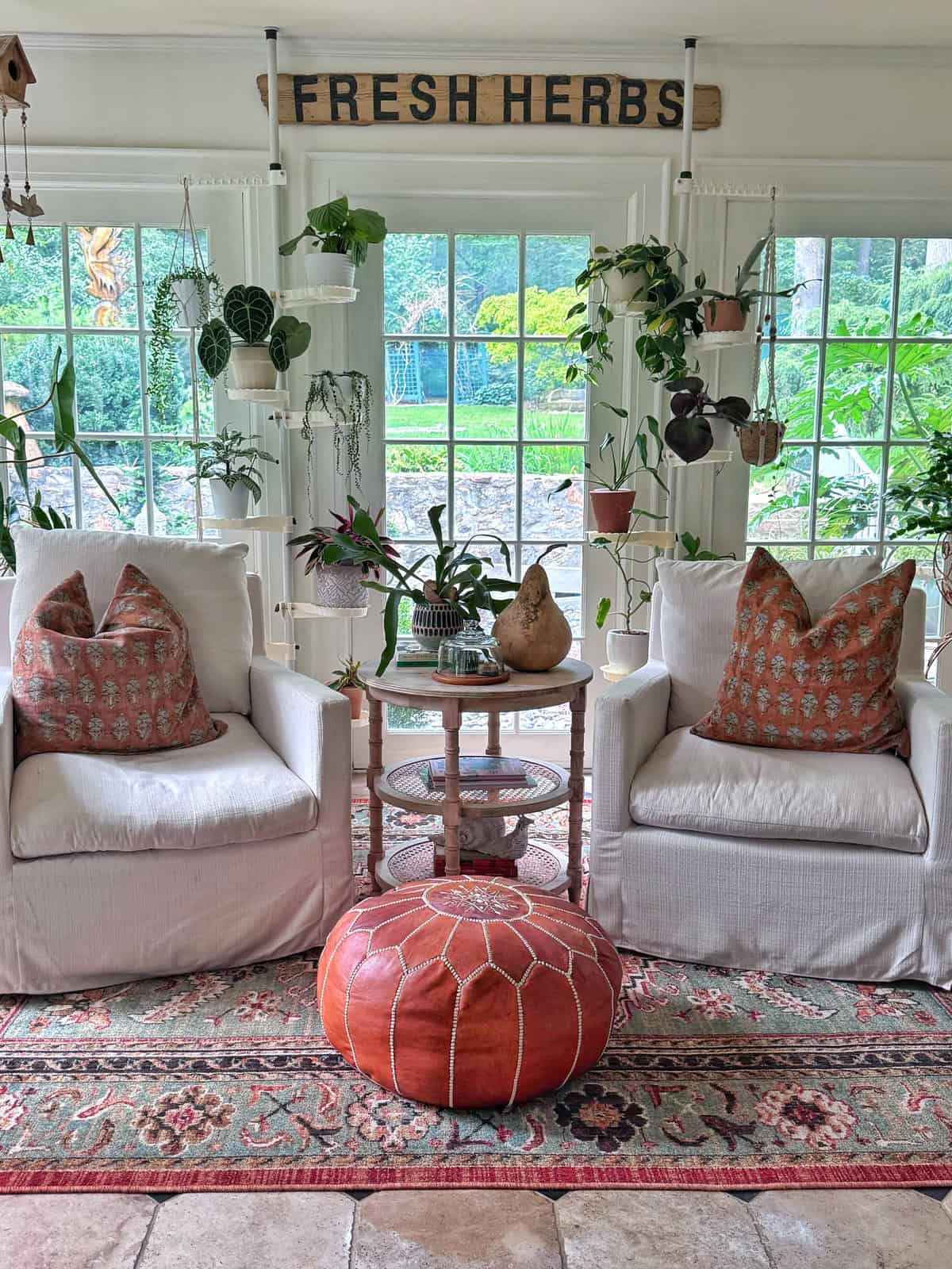 Two white armchairs with patterned pillows sit by a window with hanging plants. A round orange pouf is in front, a small table with decor between chairs, and a “FRESH HERBS” sign hangs above.