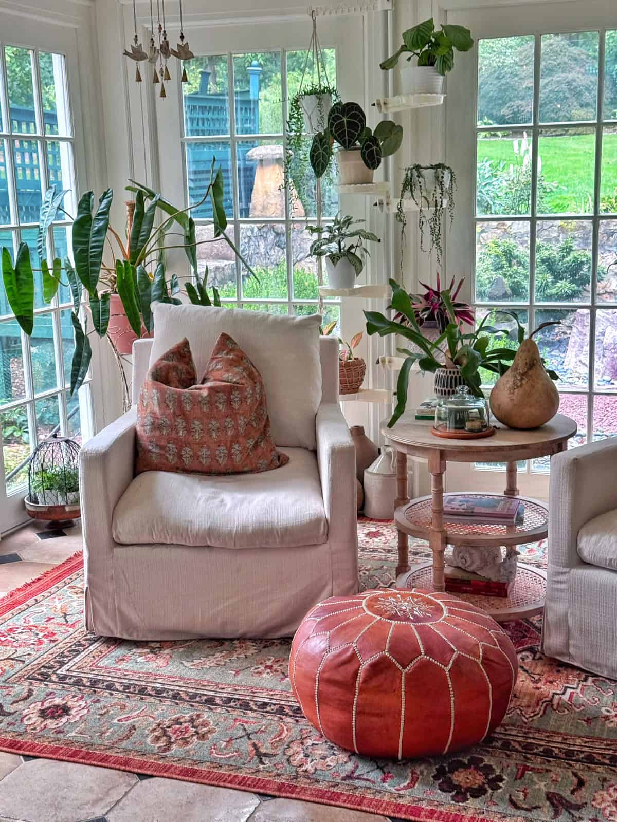 Bright sunroom with white armchairs, patterned pillows, a round orange ottoman, hanging and potted plants, a round side table, and large windows offering a green garden view.